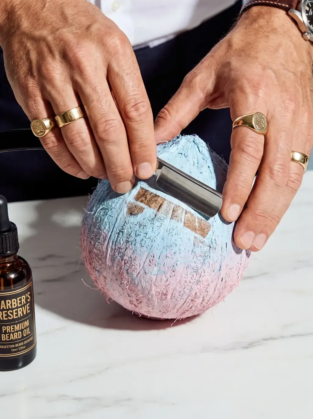 Mains ornées de bagues appliquant un rasoir sur une noix de coco peinte en pastel, flacon d'huile à barbe Barber's Reserve