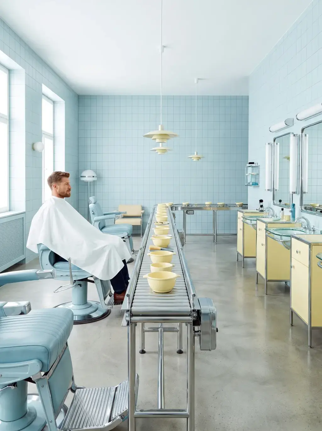 Vue d'ensemble d'un salon de coiffure carrelé bleu pastel, tapis roulant de bols jaunes entre fauteuils vintage et miroirs