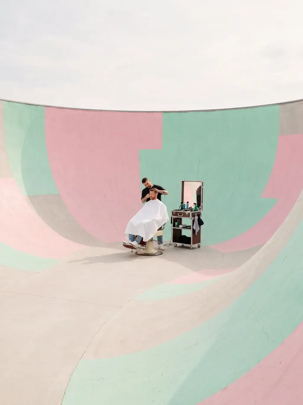 Coiffeur en pleine coupe au centre d'un skatepark aux courbes peintes en pastel rose et menthe, vue en plongée