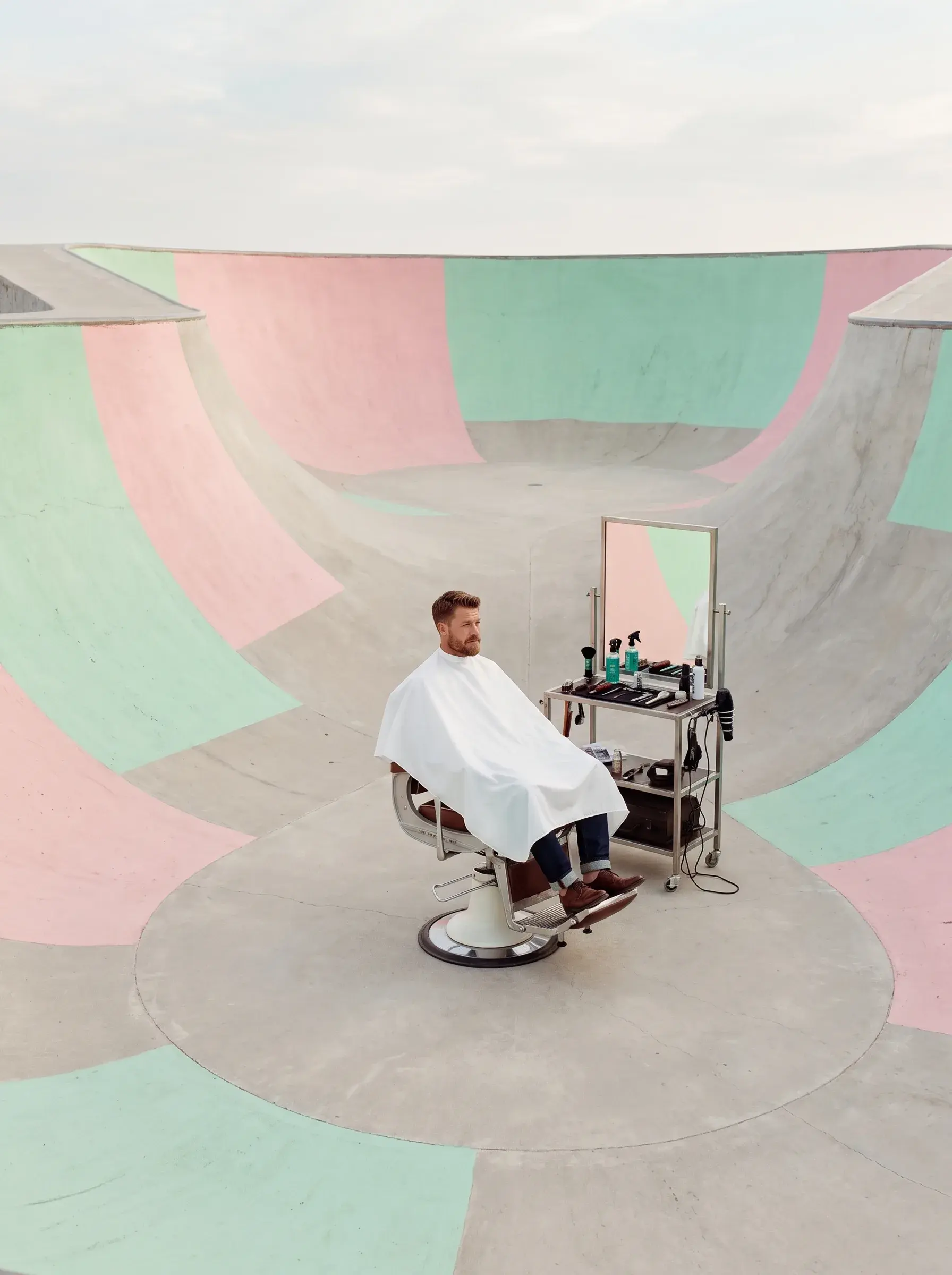 Homme en cape assis dans un fauteuil de barbier vintage au centre d'un skatepark pastel rose et vert, vue aérienne