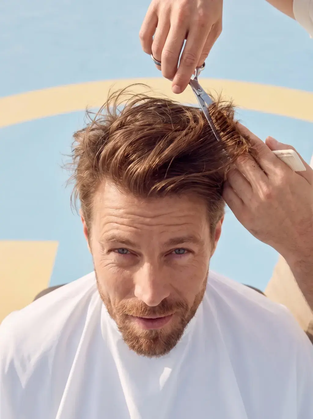 Portrait d'un homme se faisant couper les cheveux en extérieur, fond pastel bleu et jaune
