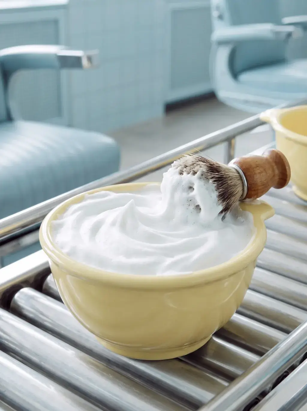 Blaireau et mousse à raser dans un bol jaune posés sur un chariot de barbier chromé, salon pastel bleu en arrière-plan