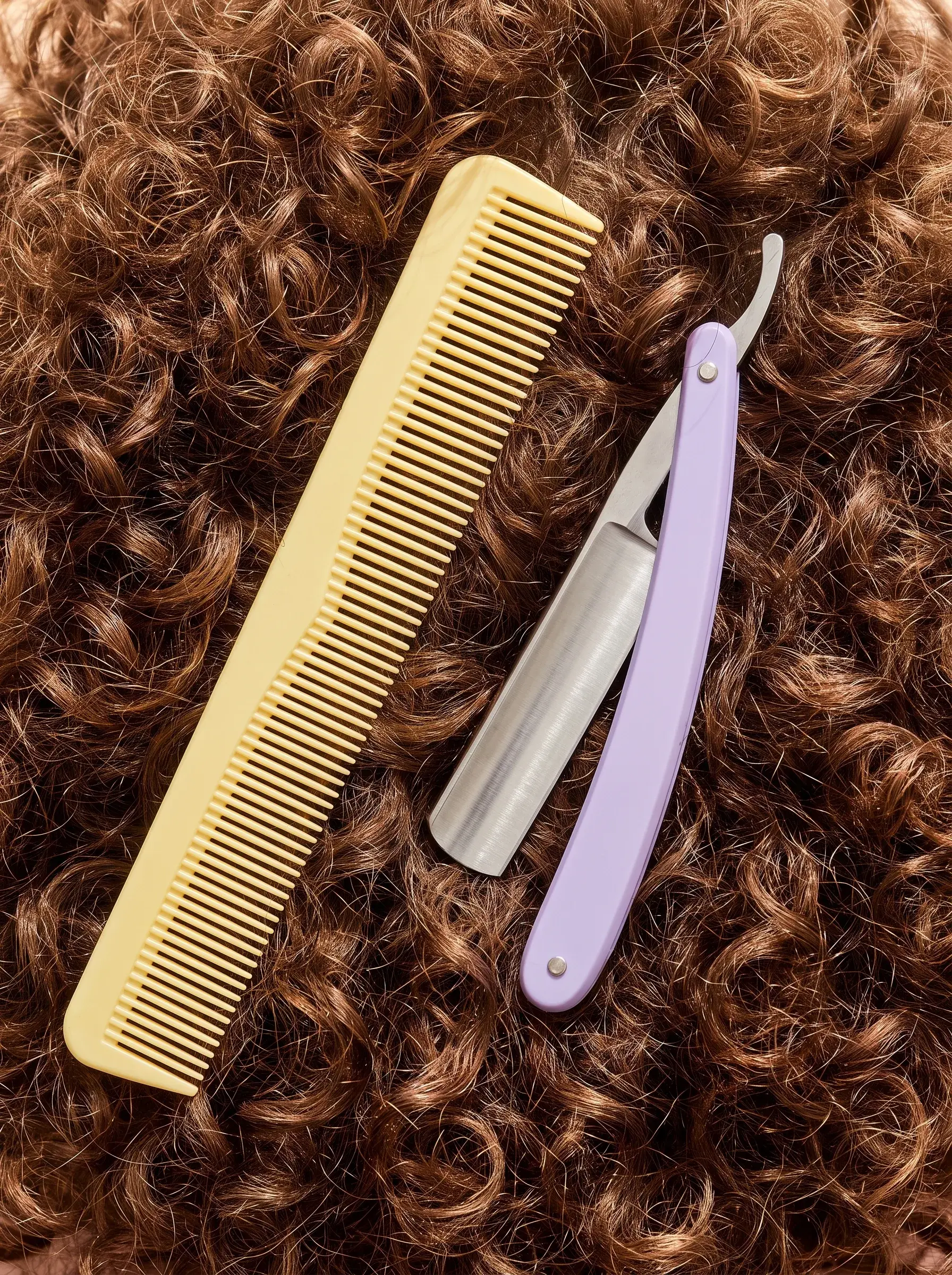 Peigne jaune et rasoir coupe-chou lilas posés sur une chevelure brune bouclée