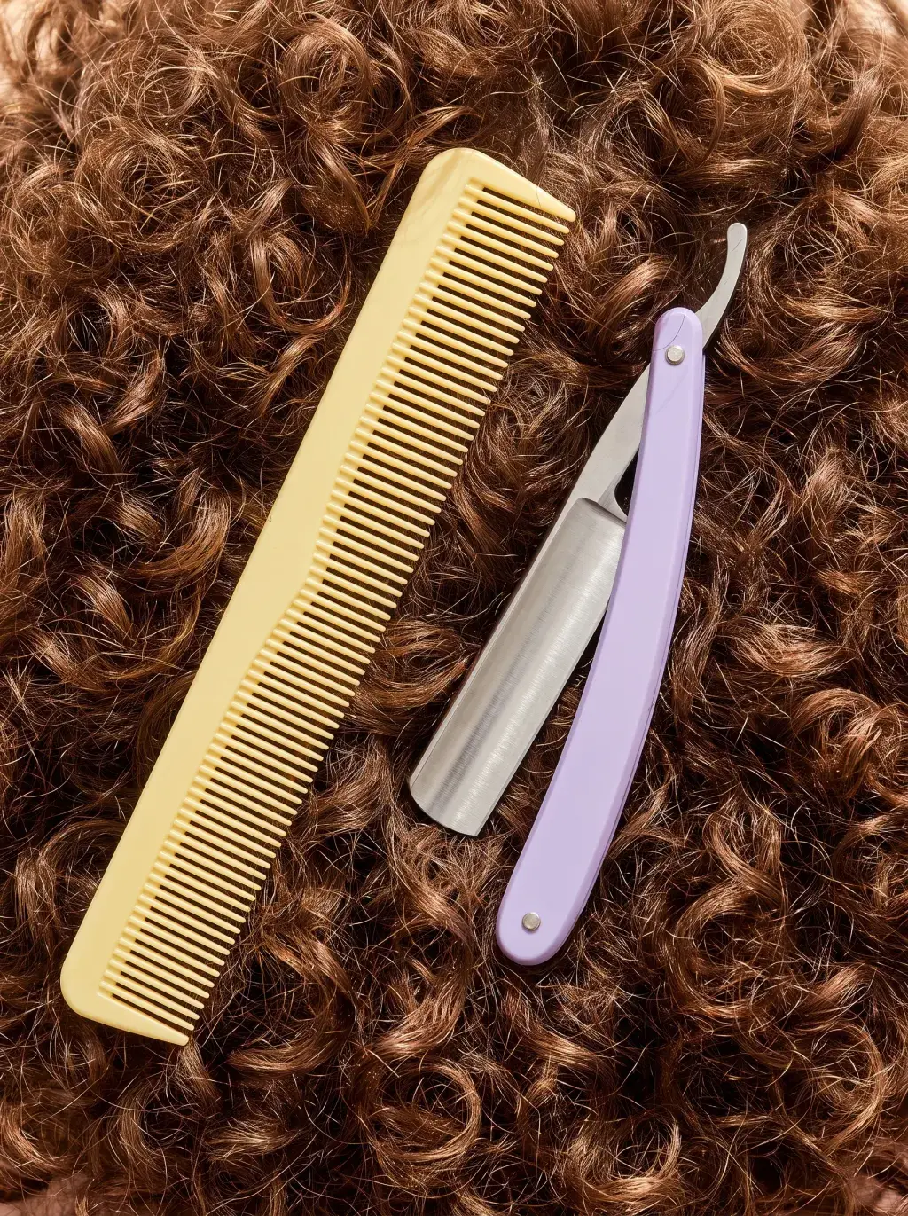 Peigne jaune et rasoir coupe-chou lilas posés sur une chevelure brune bouclée