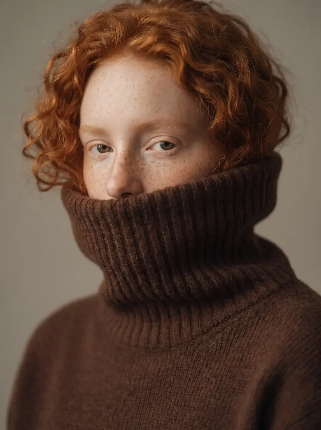 Portrait d'une jeune femme rousse le visage enfoui dans le col roulé d'un pull marron épais