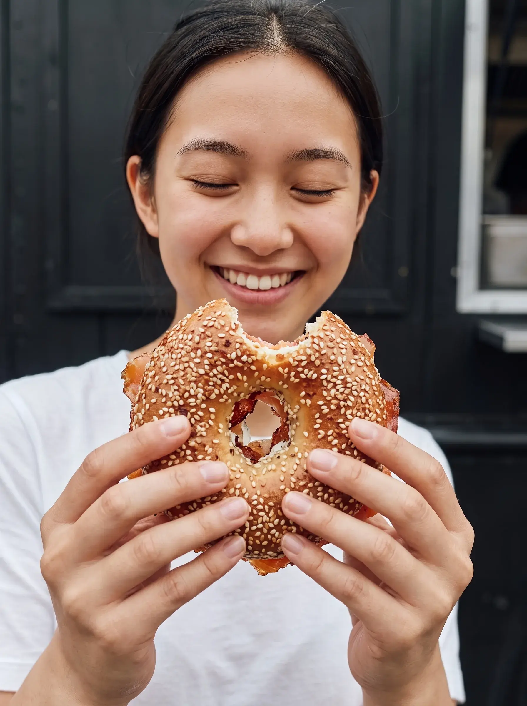 Jeune femme asiatique souriante tenant un bagel mordu devant une porte noire