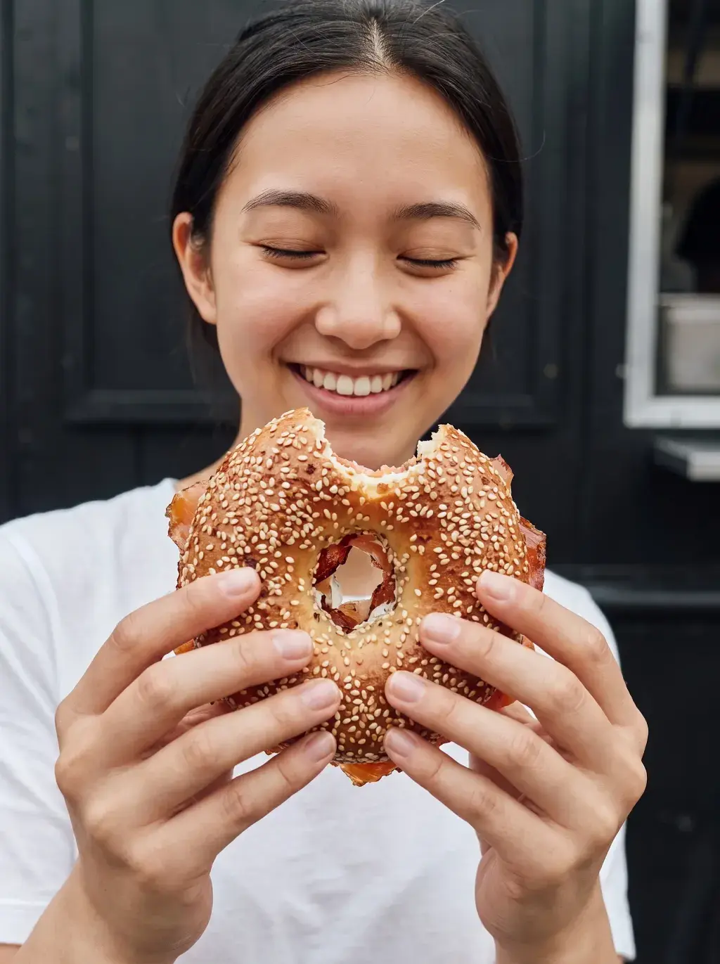 Jeune femme asiatique souriante tenant un bagel mordu devant une porte noire