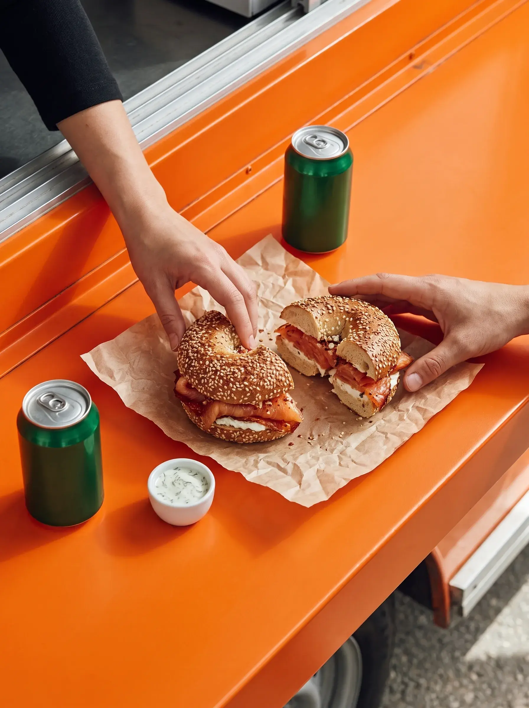 Vue plongeante sur deux mains partageant un bagel saumon sur comptoir orange avec canettes vertes