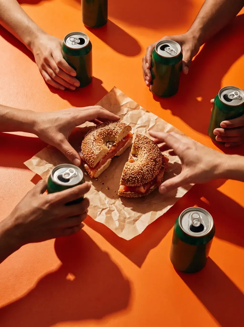 Vue plongeante sur plusieurs mains attrapant un bagel partagé entre canettes vertes sur fond orange