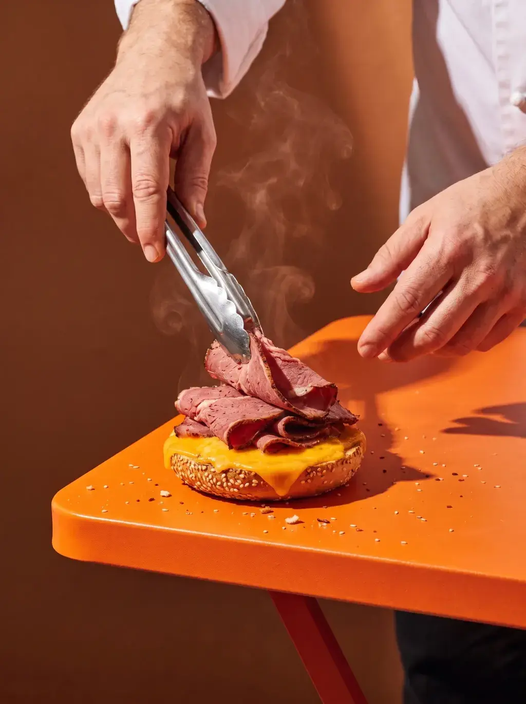 Chef déposant des tranches de pastrami fumantes avec une pince sur un bagel cheese sur table orange
