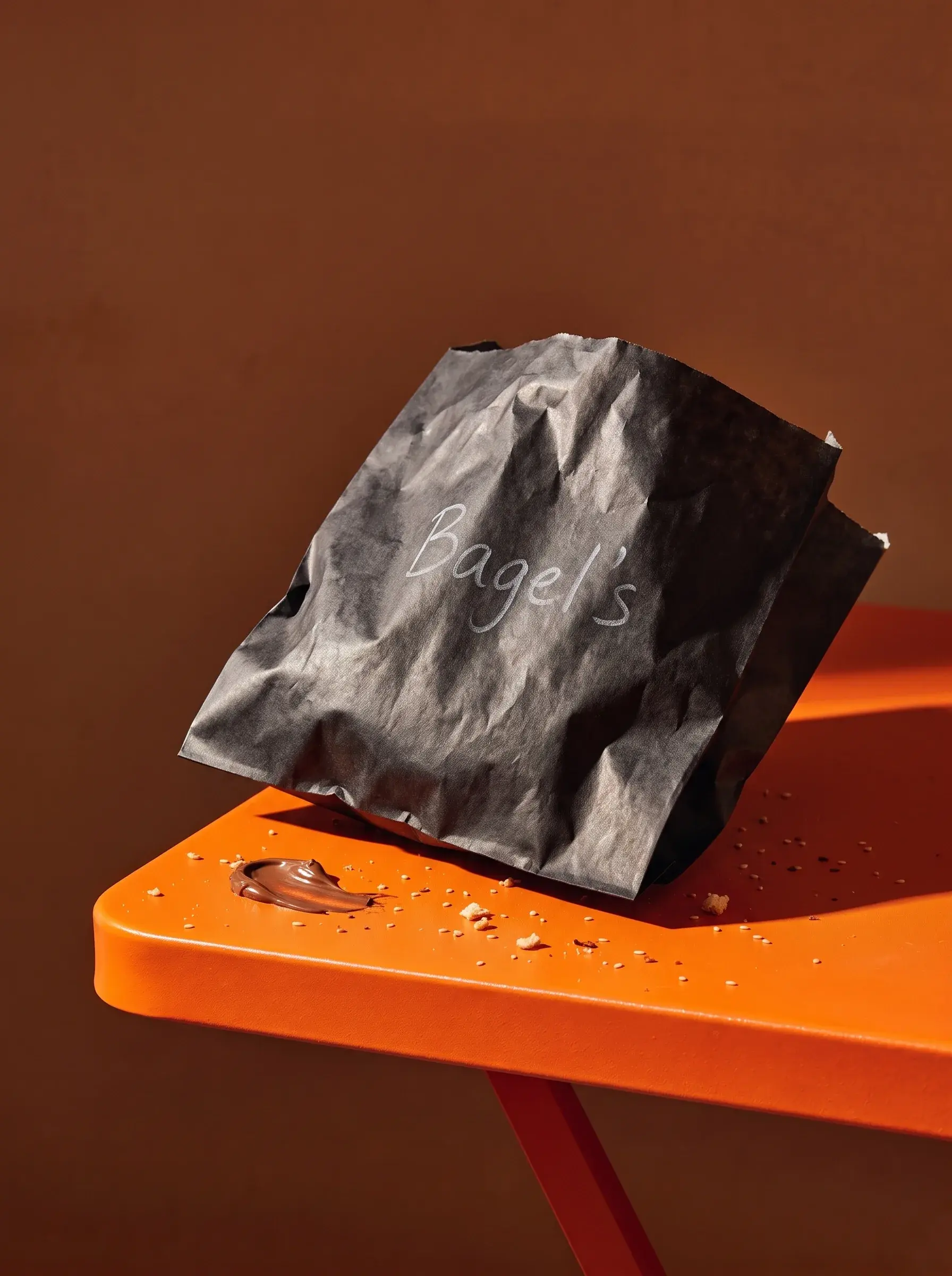 Sac papier noir Bagel's froissé posé sur une table orange avec miettes et trace de sauce