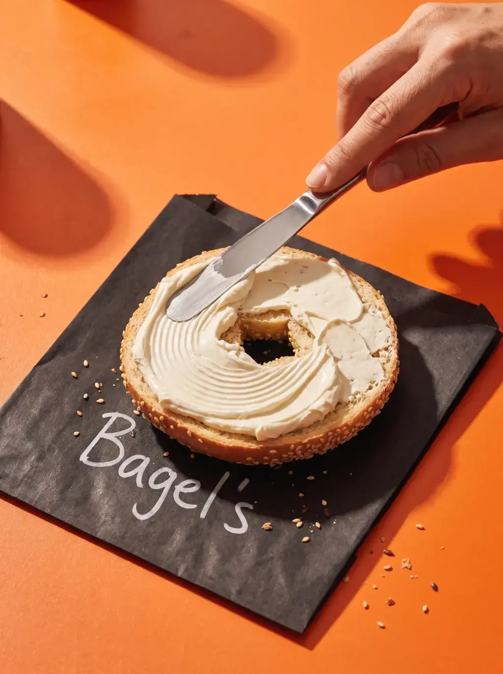 Main étalant du cream cheese avec un couteau sur un bagel posé sur papier Bagel's fond orange