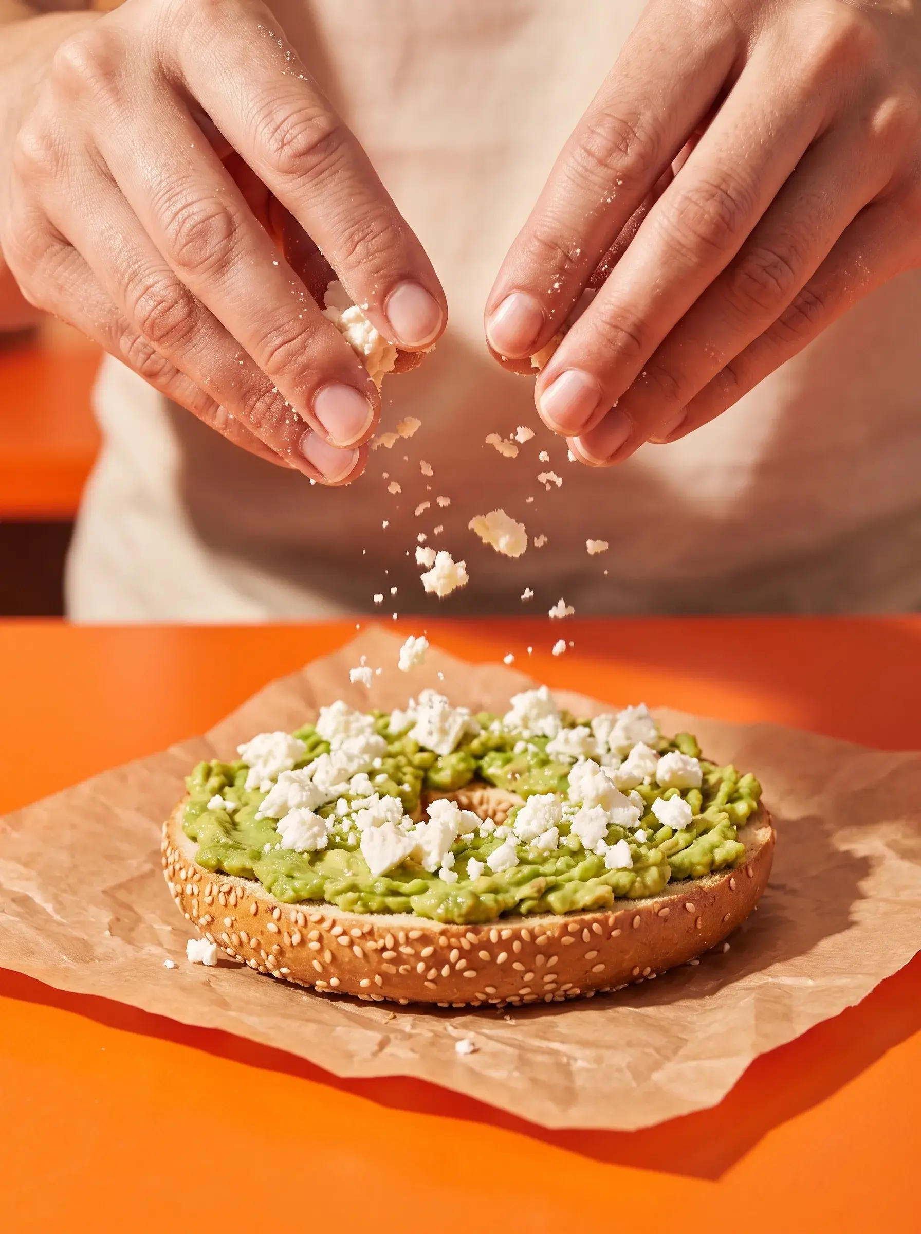 Mains émiettant de la feta sur un bagel avocat écrasé sur papier kraft fond orange