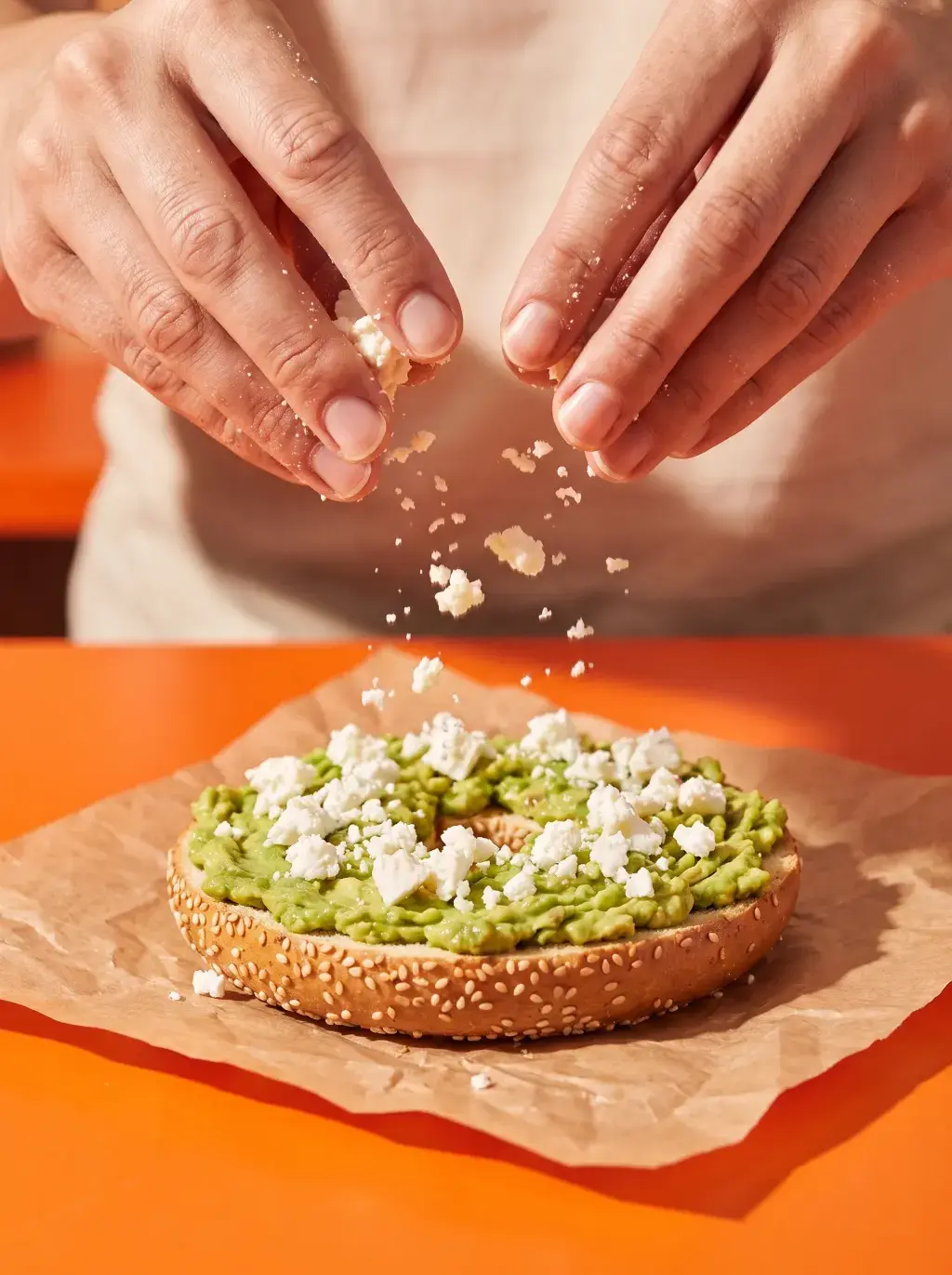 Mains émiettant de la feta sur un bagel avocat écrasé sur papier kraft fond orange