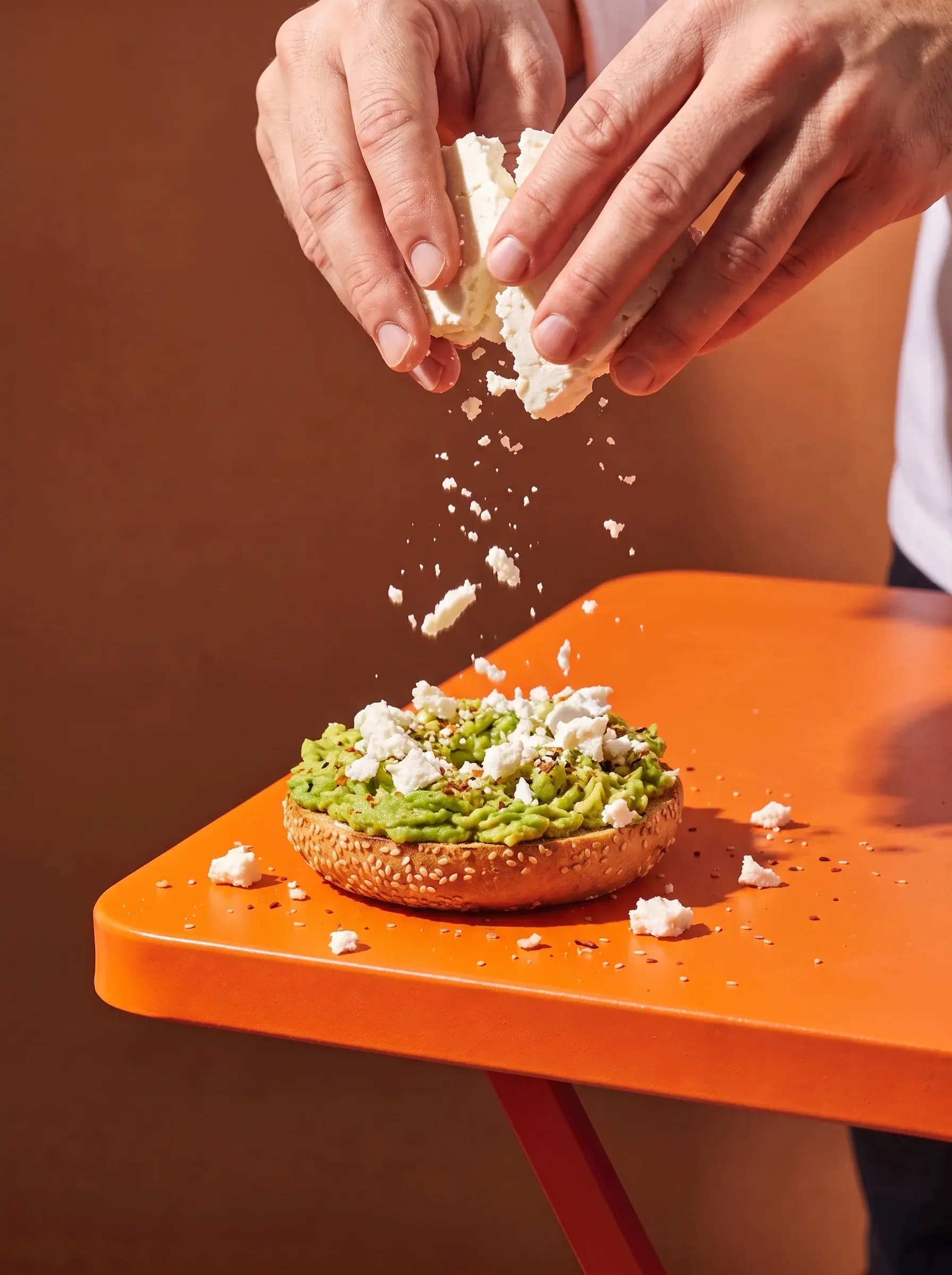 Mains émiettant de la feta sur un bagel avocat posé sur une table orange avec épices