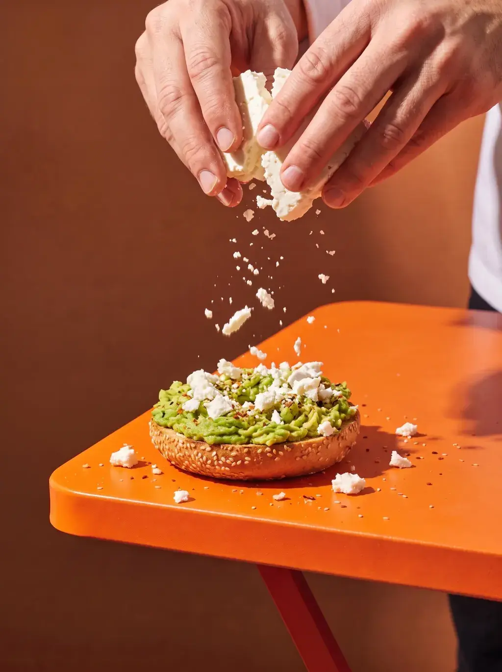 Mains émiettant de la feta sur un bagel avocat posé sur une table orange avec épices