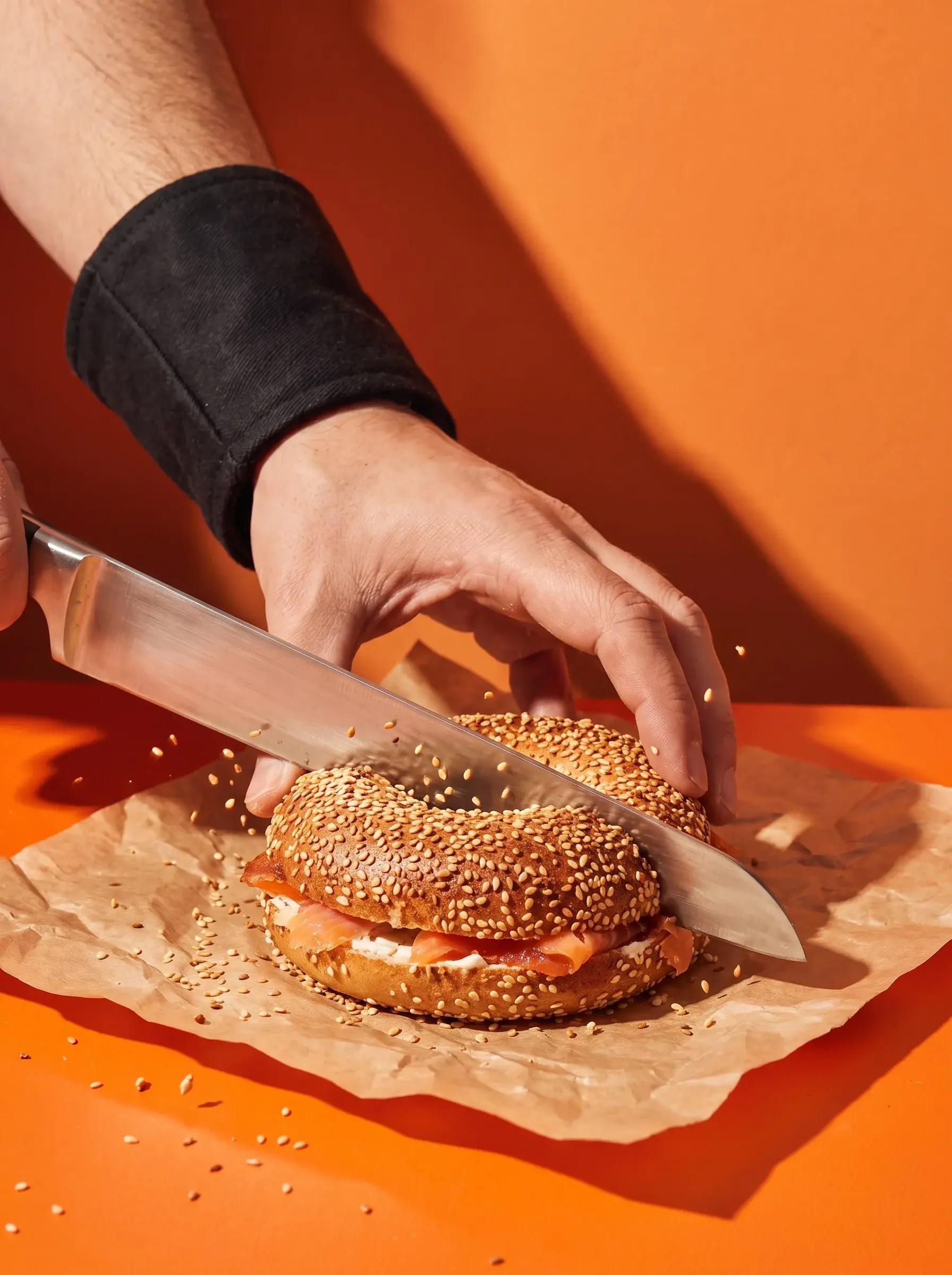 Main avec bracelet noir coupant un bagel saumon cream cheese avec un grand couteau sur fond orange