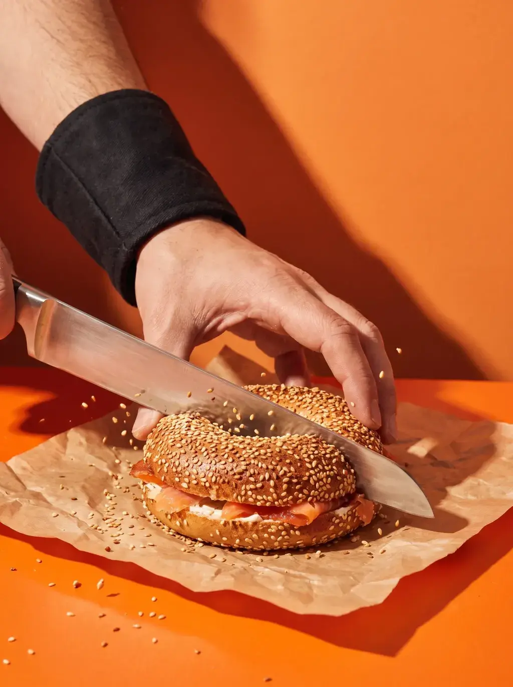 Main avec bracelet noir coupant un bagel saumon cream cheese avec un grand couteau sur fond orange