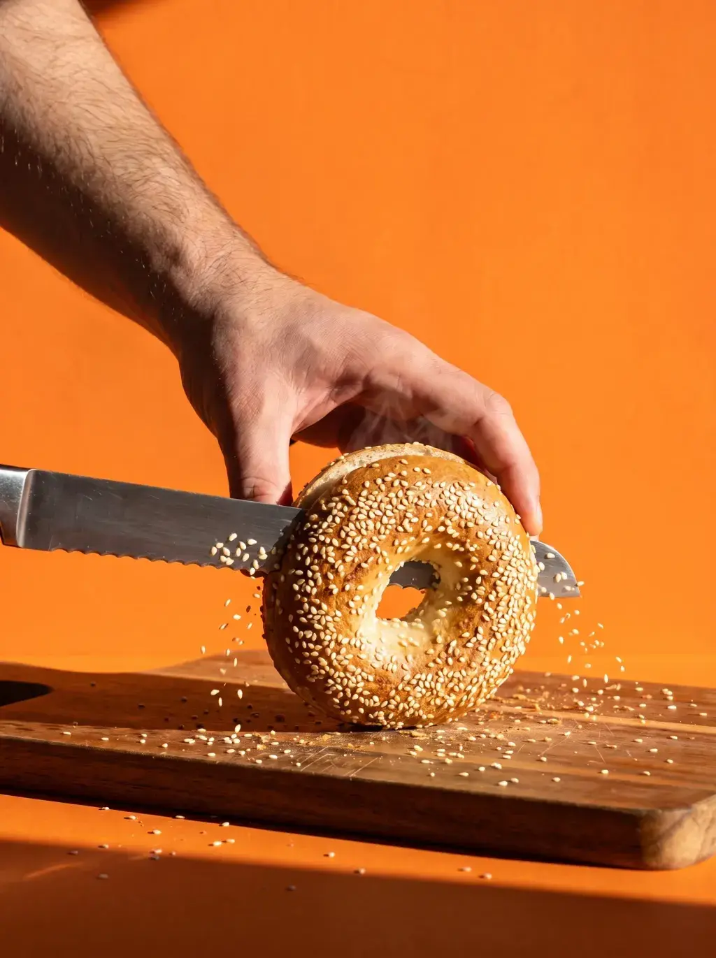 Main coupant un bagel au sésame avec un couteau à pain sur planche bois graines qui volent fond orange