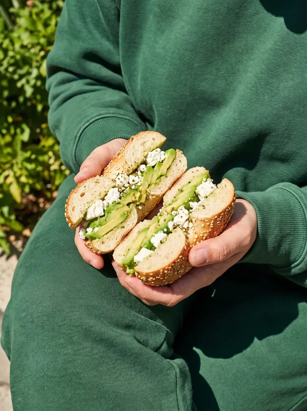 Mains en sweat vert tenant un bagel avocat feta coupé en deux sous lumière naturelle extérieure