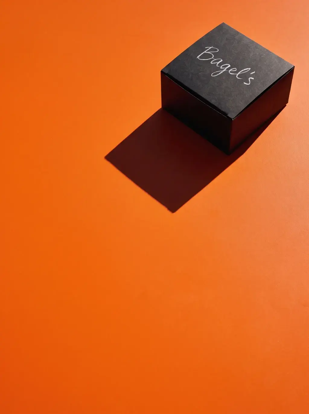 Boîte noire carrée Bagel's fermée sur fond orange avec longue ombre portée graphique