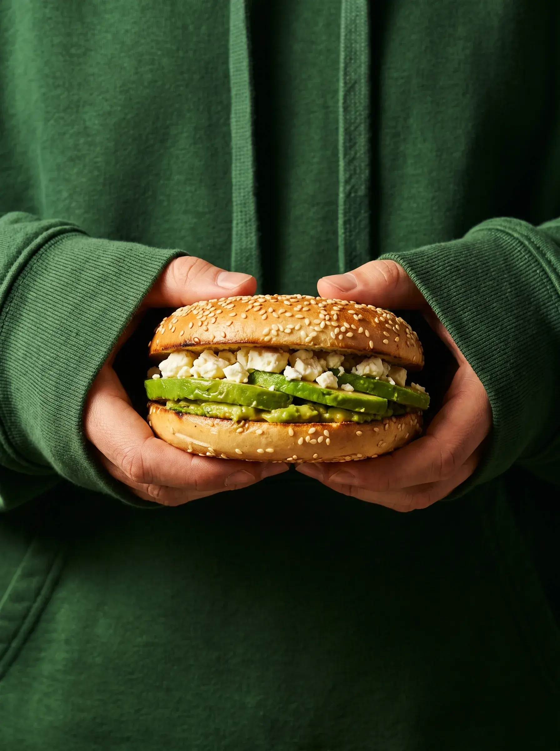 Mains en sweat vert tenant un bagel avocat feta sur fond vert sombre composition monochrome