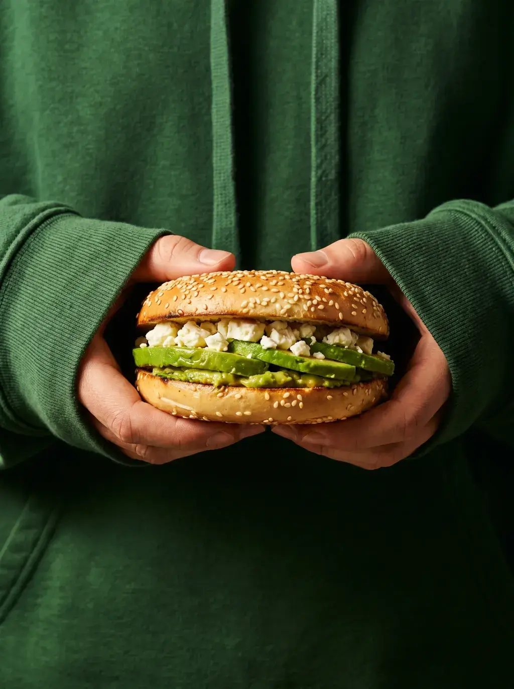 Mains en sweat vert tenant un bagel avocat feta sur fond vert sombre composition monochrome