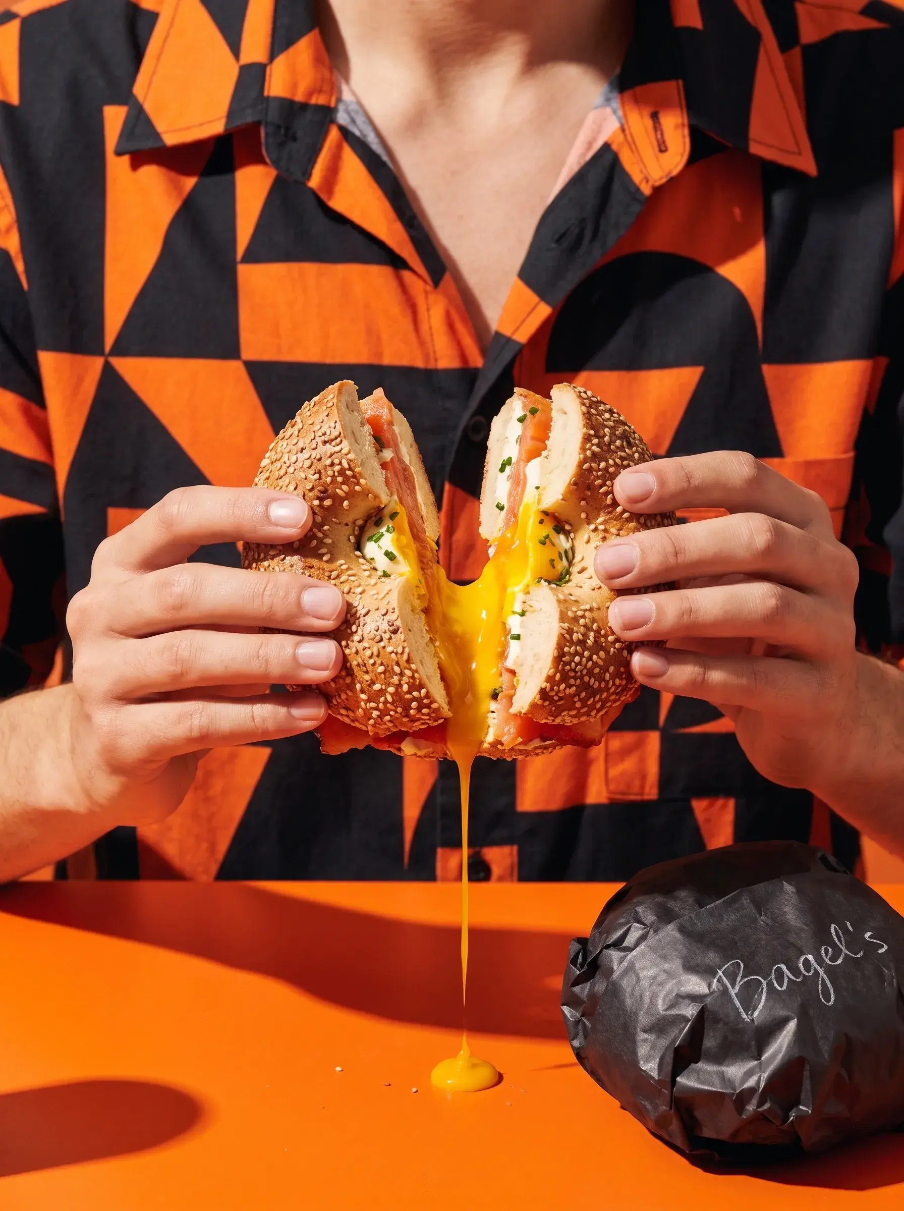 Personne en chemise orange et noir ouvrant un bagel saumon œuf coulant sur fond orange avec sac Bagel's