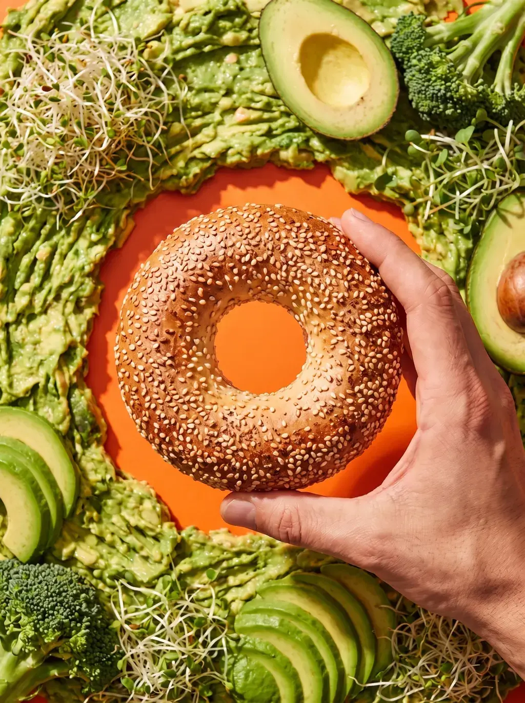 Main tenant un bagel au sésame au-dessus d'un lit d'ingrédients verts sur fond orange