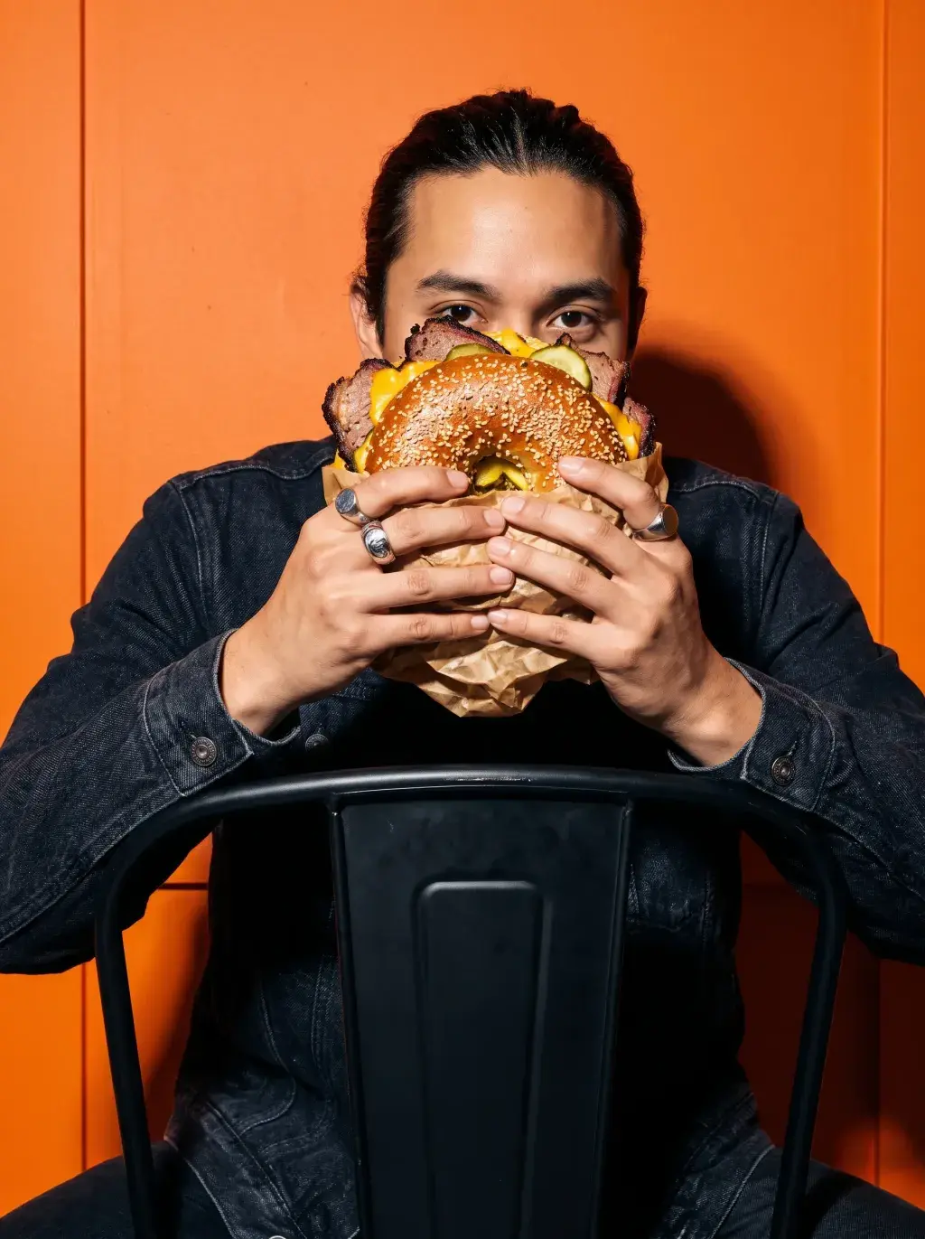 Homme cachant son visage derrière un énorme bagel brisket cornichons sur fond orange