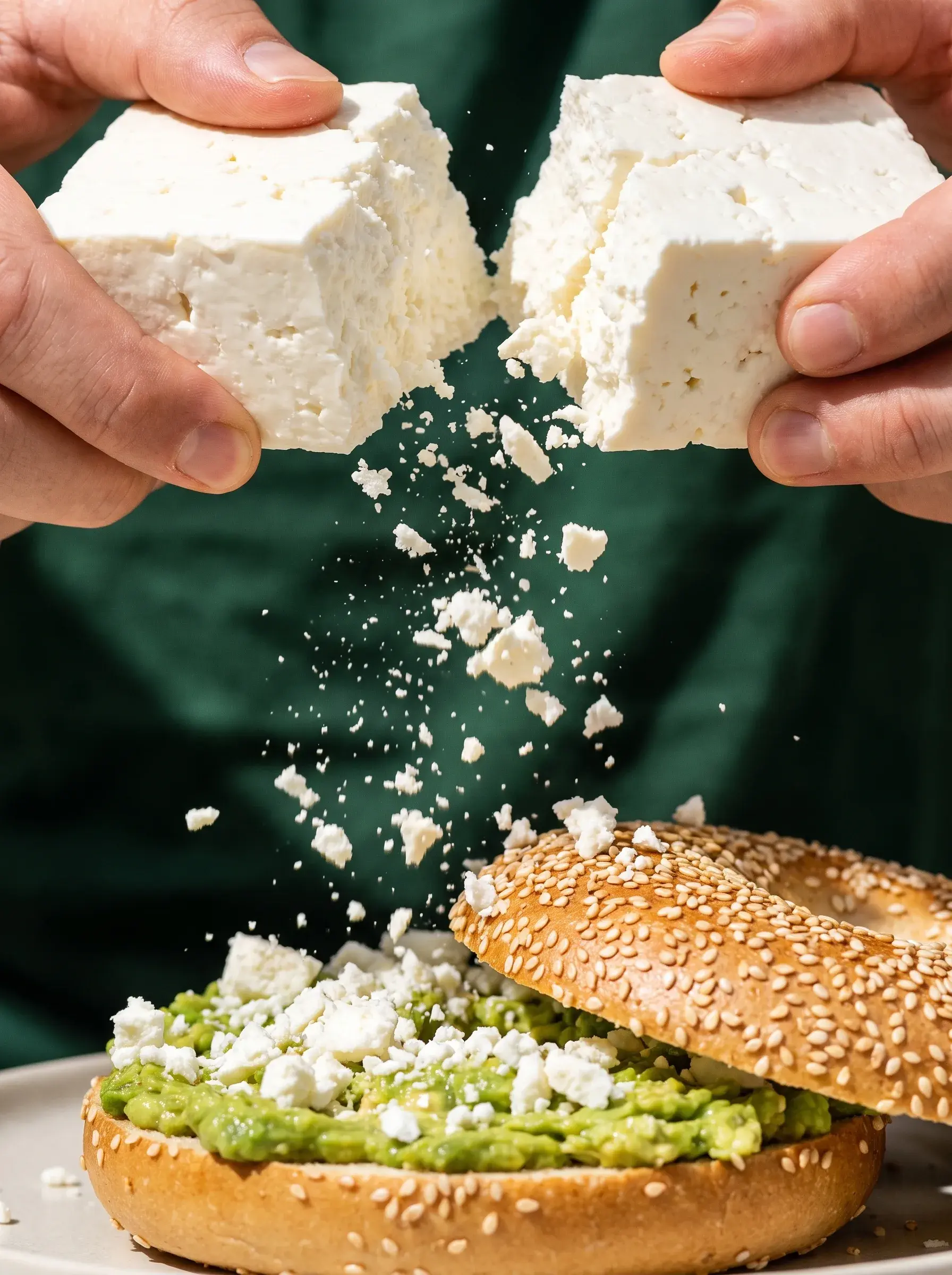 Mains cassant un bloc de feta au-dessus d'un bagel avocat ouvert sur fond vert