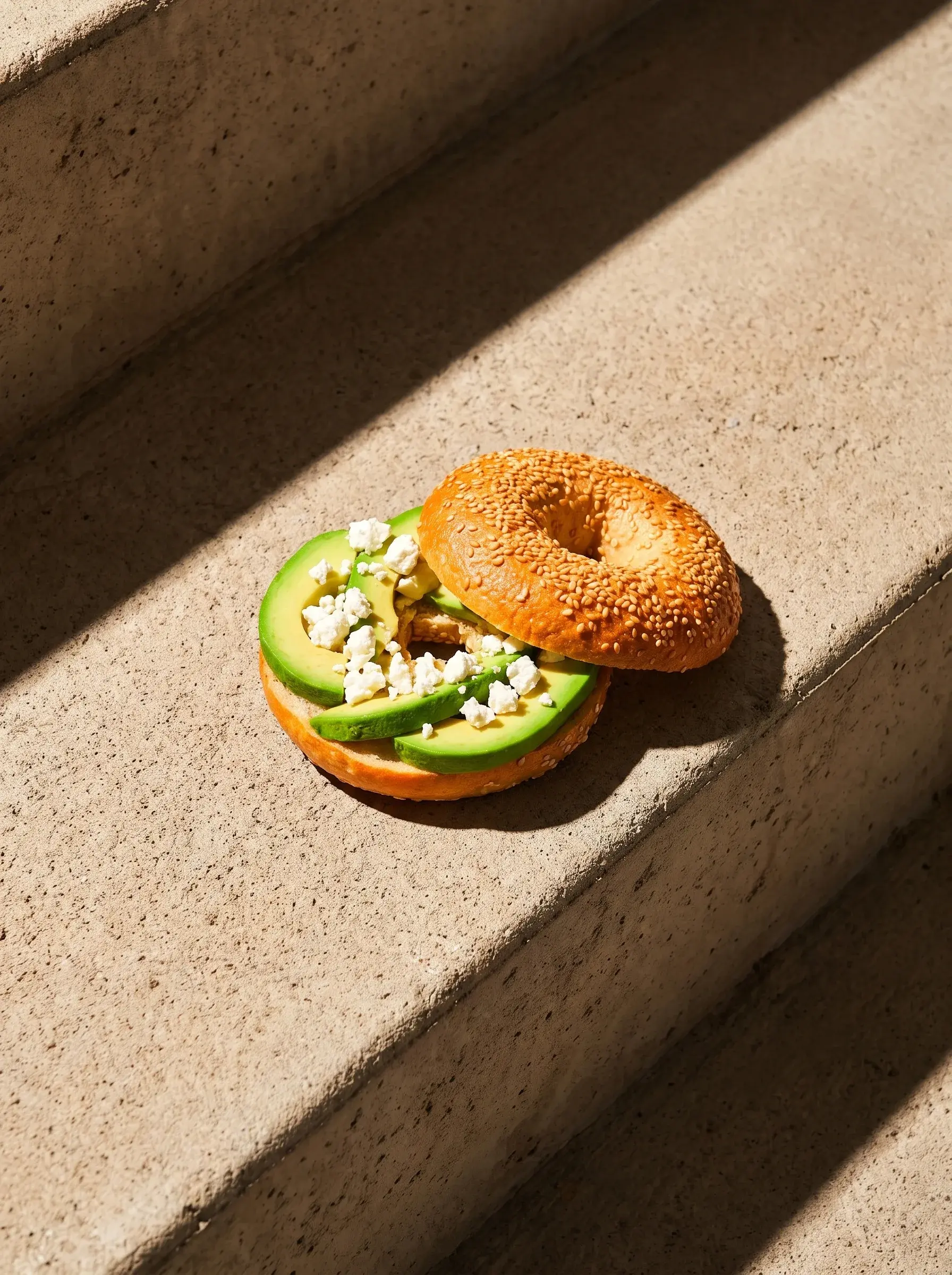 Bagel avocat feta ouvert sur papier kraft sur une marche en pierre sous lumière rasante