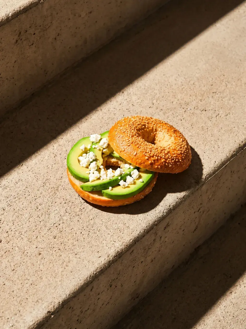 Bagel avocat feta ouvert sur papier kraft sur une marche en pierre sous lumière rasante