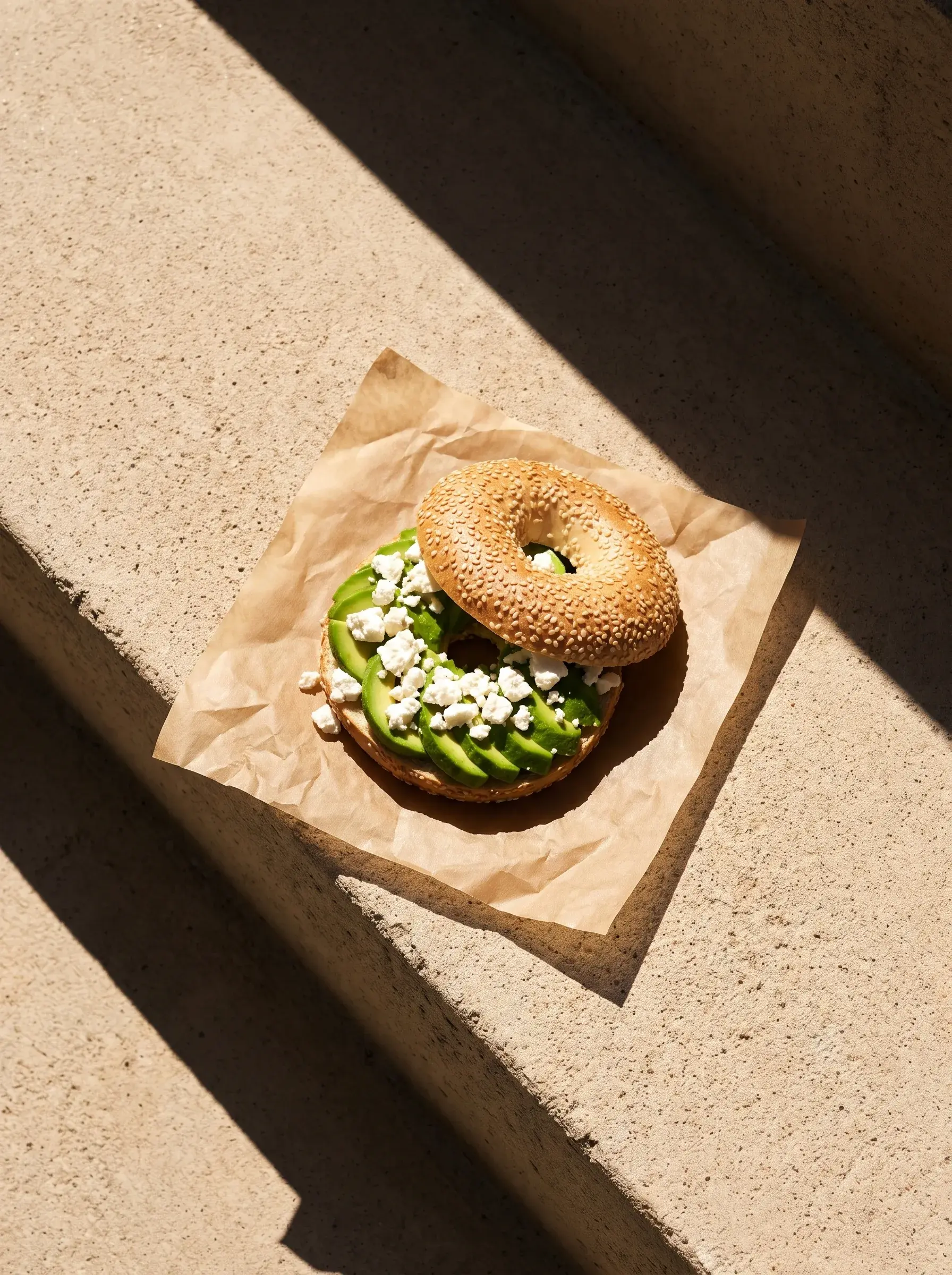 Bagel avocat feta sur papier kraft en angle sur une marche de pierre avec ombres diagonales