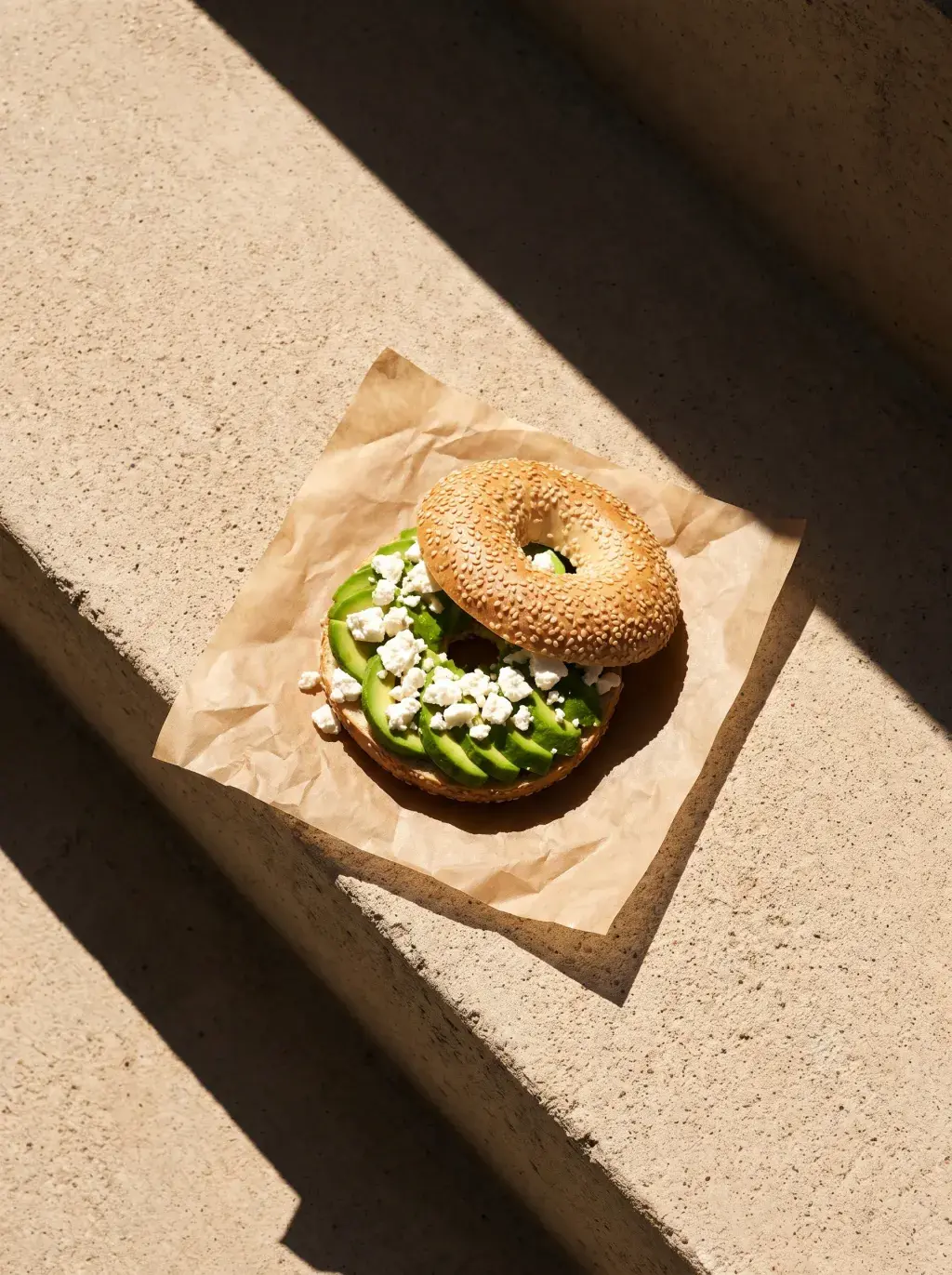 Bagel avocat feta sur papier kraft en angle sur une marche de pierre avec ombres diagonales