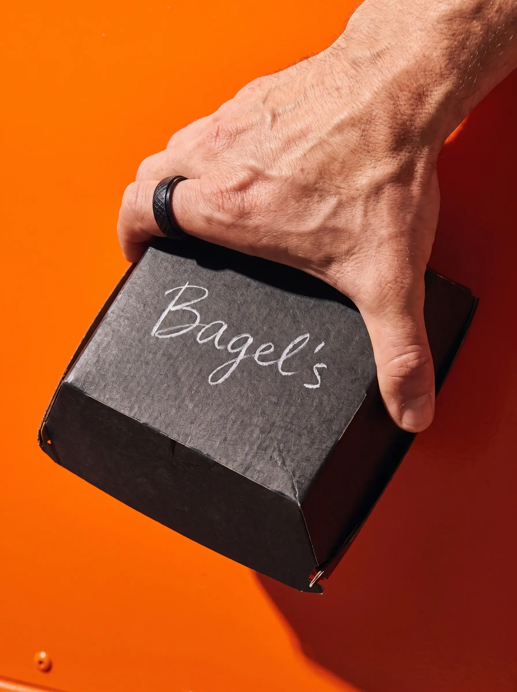 Main avec bague noire tenant une boîte Bagel's carrée sur fond orange vif