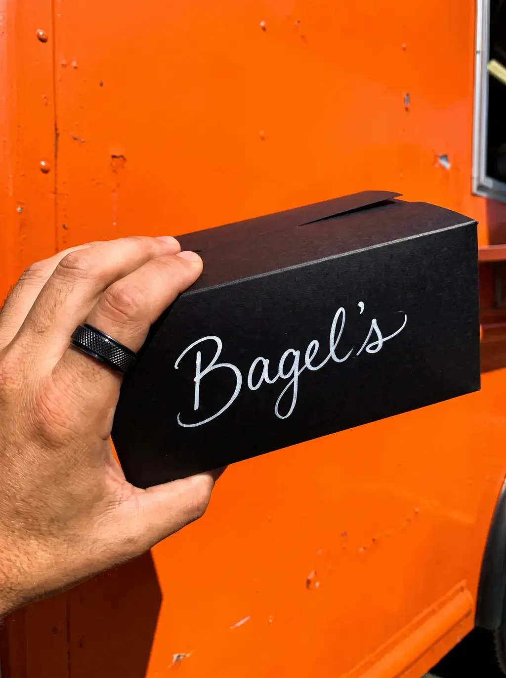 Main tenant une boîte rectangulaire Bagel's noire contre le food truck orange