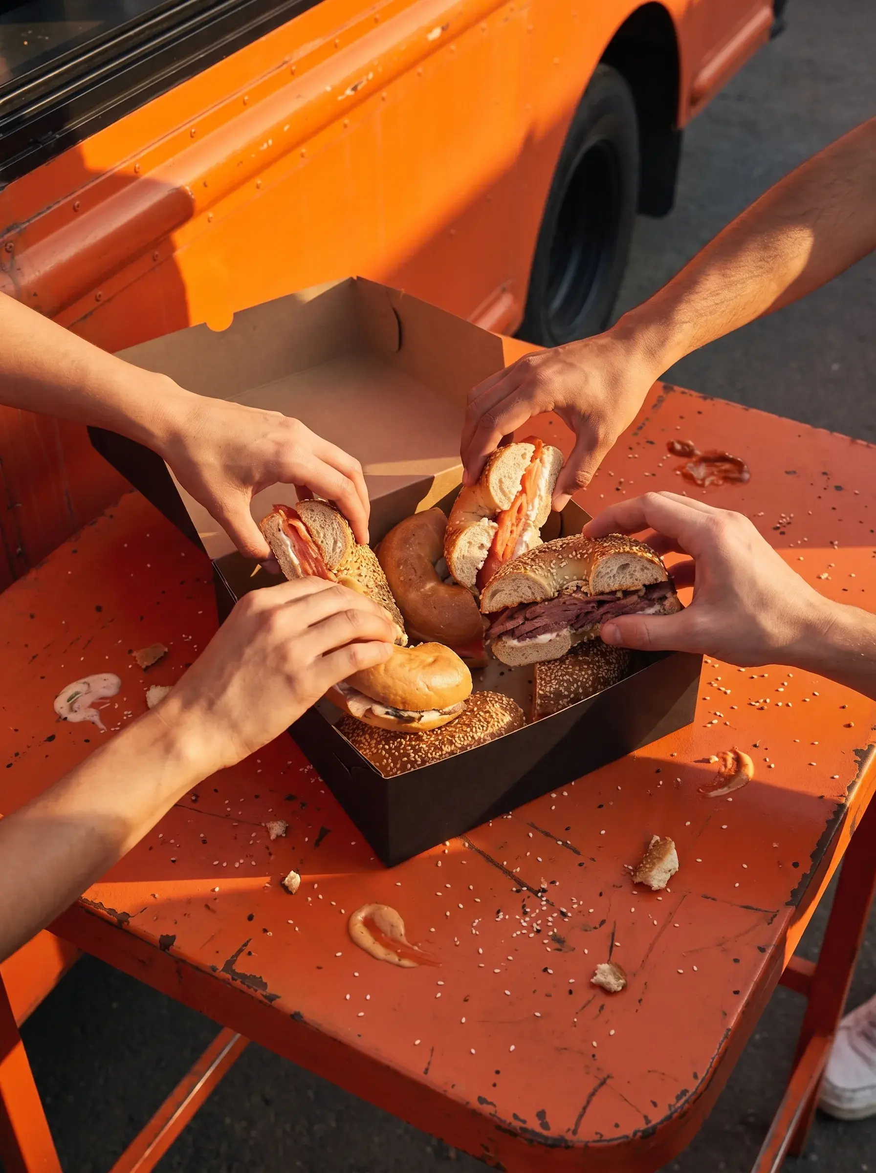 Plusieurs mains attrapant des bagels variés depuis une boîte noire sur table orange