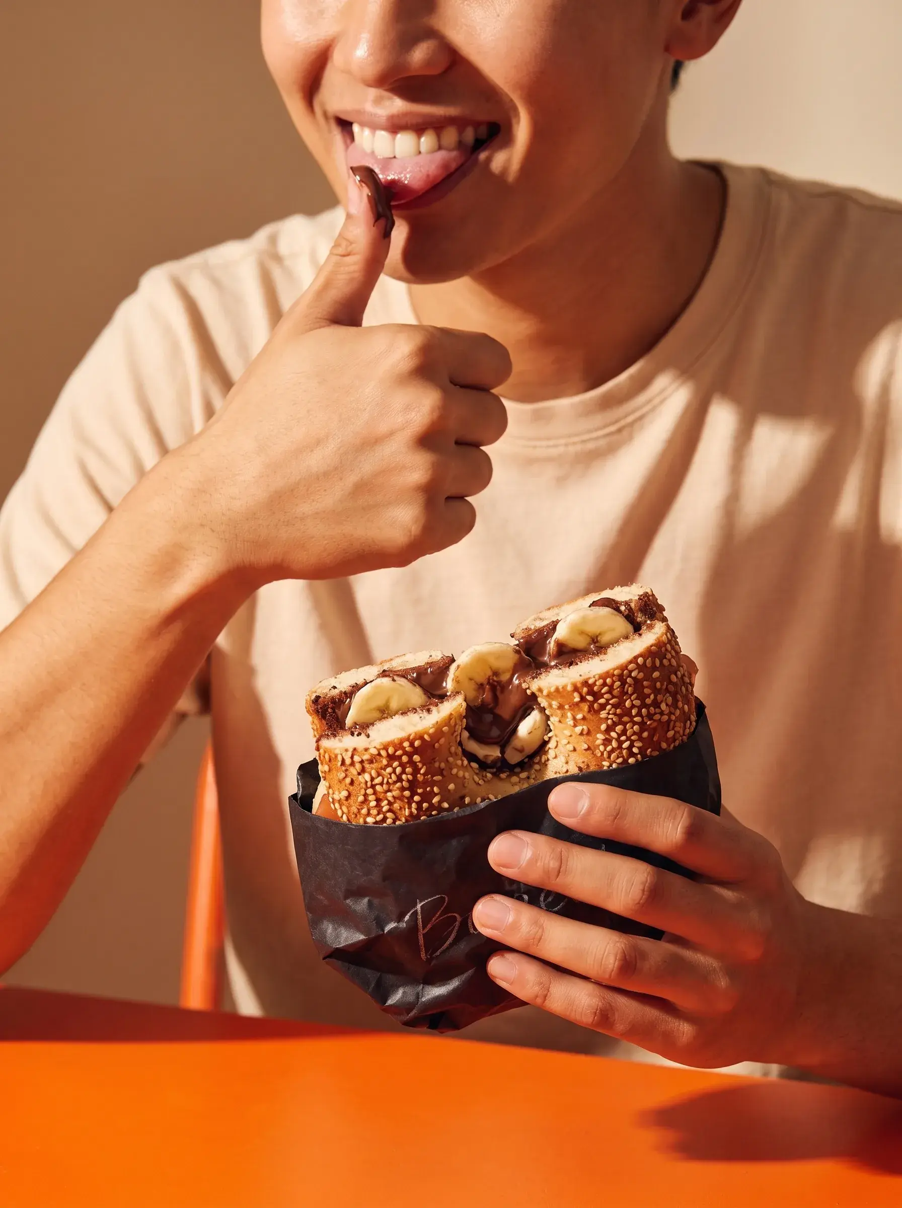 Personne léchant du chocolat sur son pouce en tenant un bagel nutella banane dans un sac Bagel's