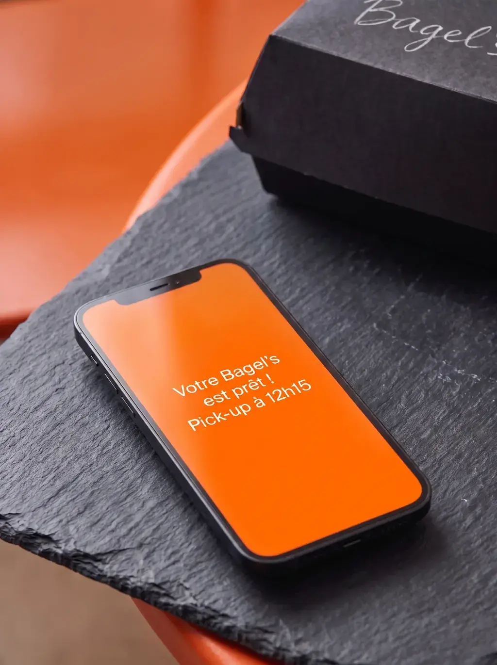 Smartphone posé sur une ardoise noire avec notification orange Bagel's prête à récupérer et boîte noire en arrière-plan