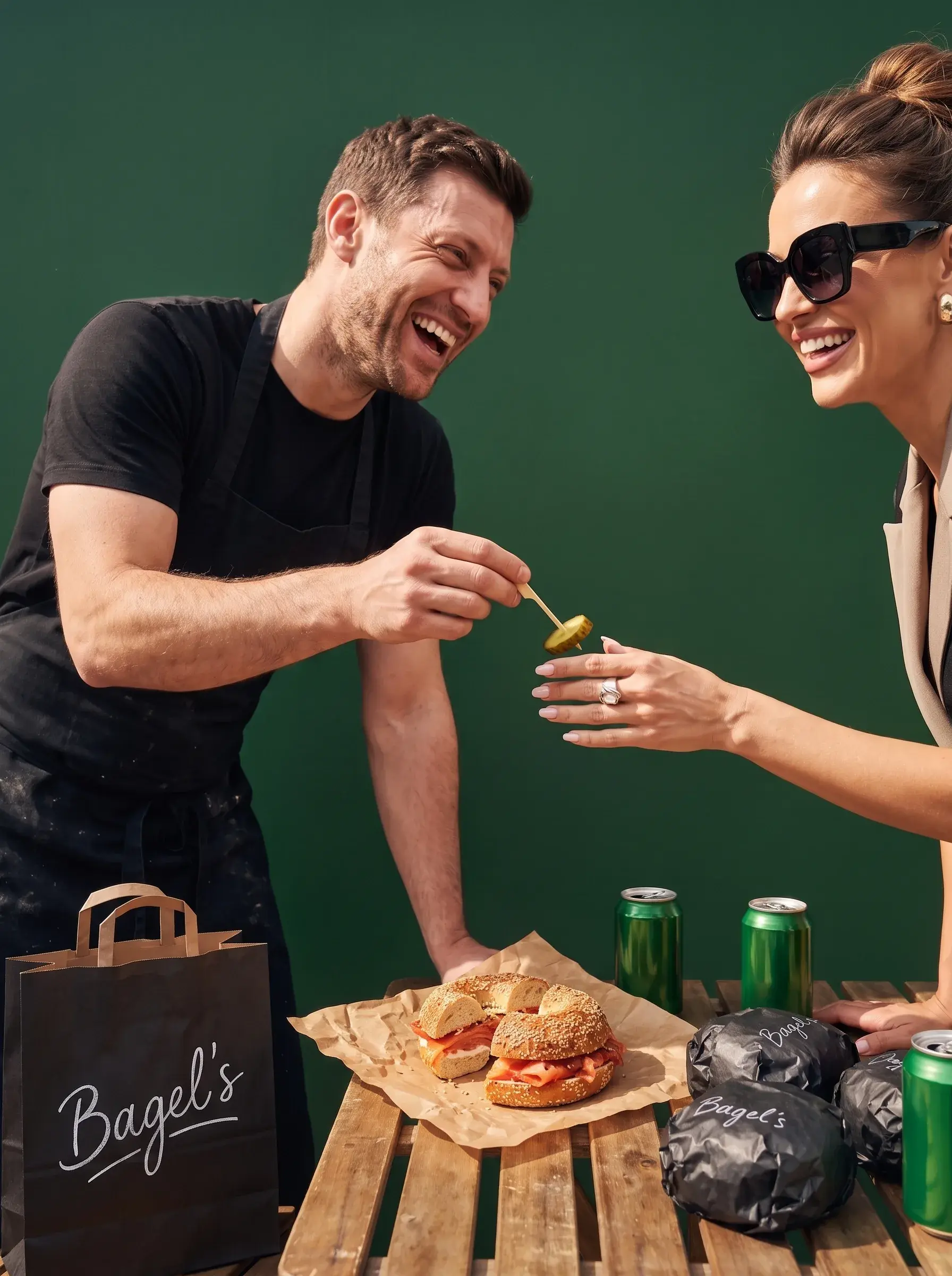 Chef en tablier noir tendant un cornichon à une femme élégante en lunettes de soleil devant bagel saumon et canettes vertes sur fond vert