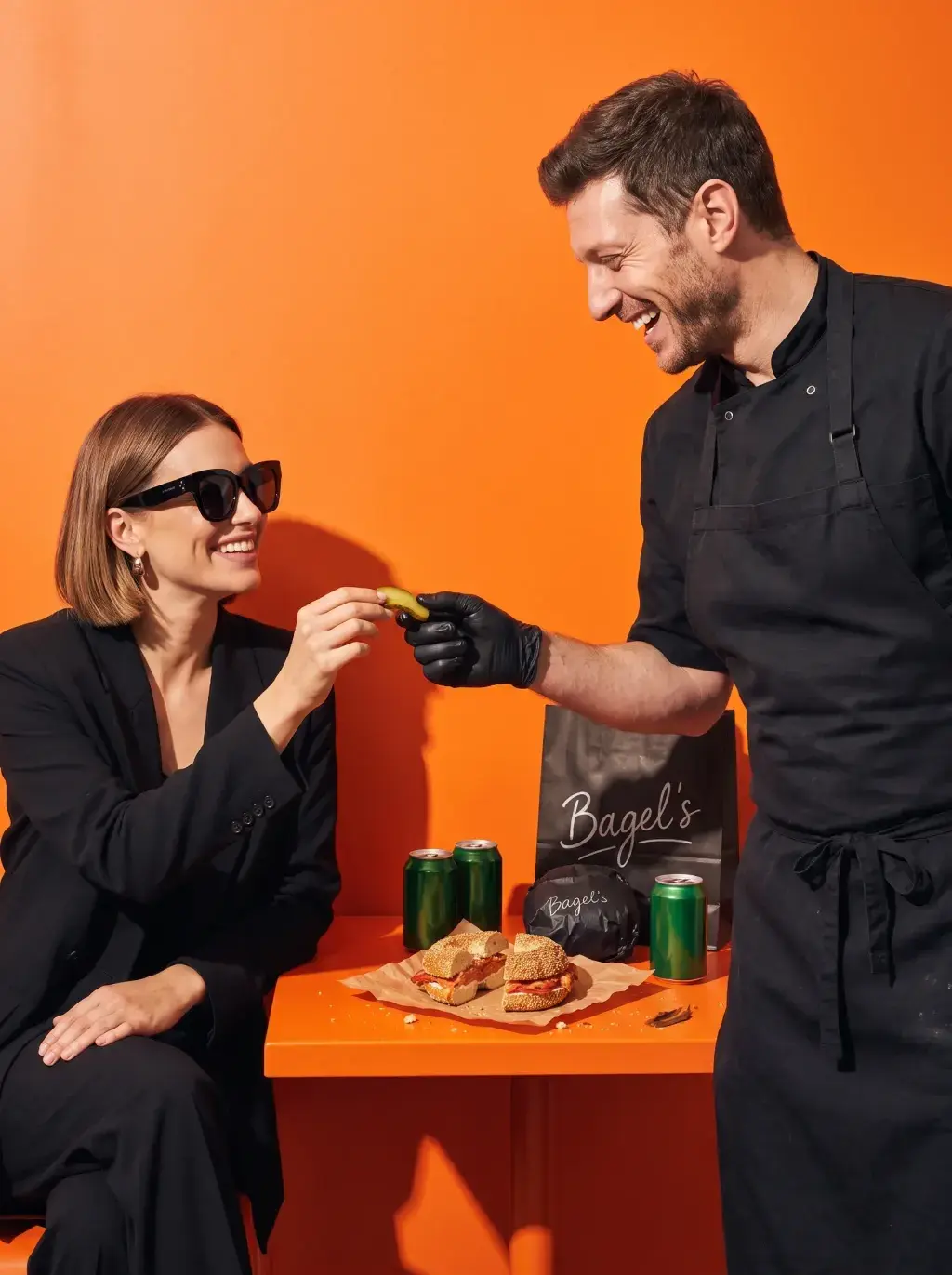 Chef en gants noirs tendant un cornichon à une cliente assise en tenue noire sur fond orange vif avec bagel et canettes vertes
