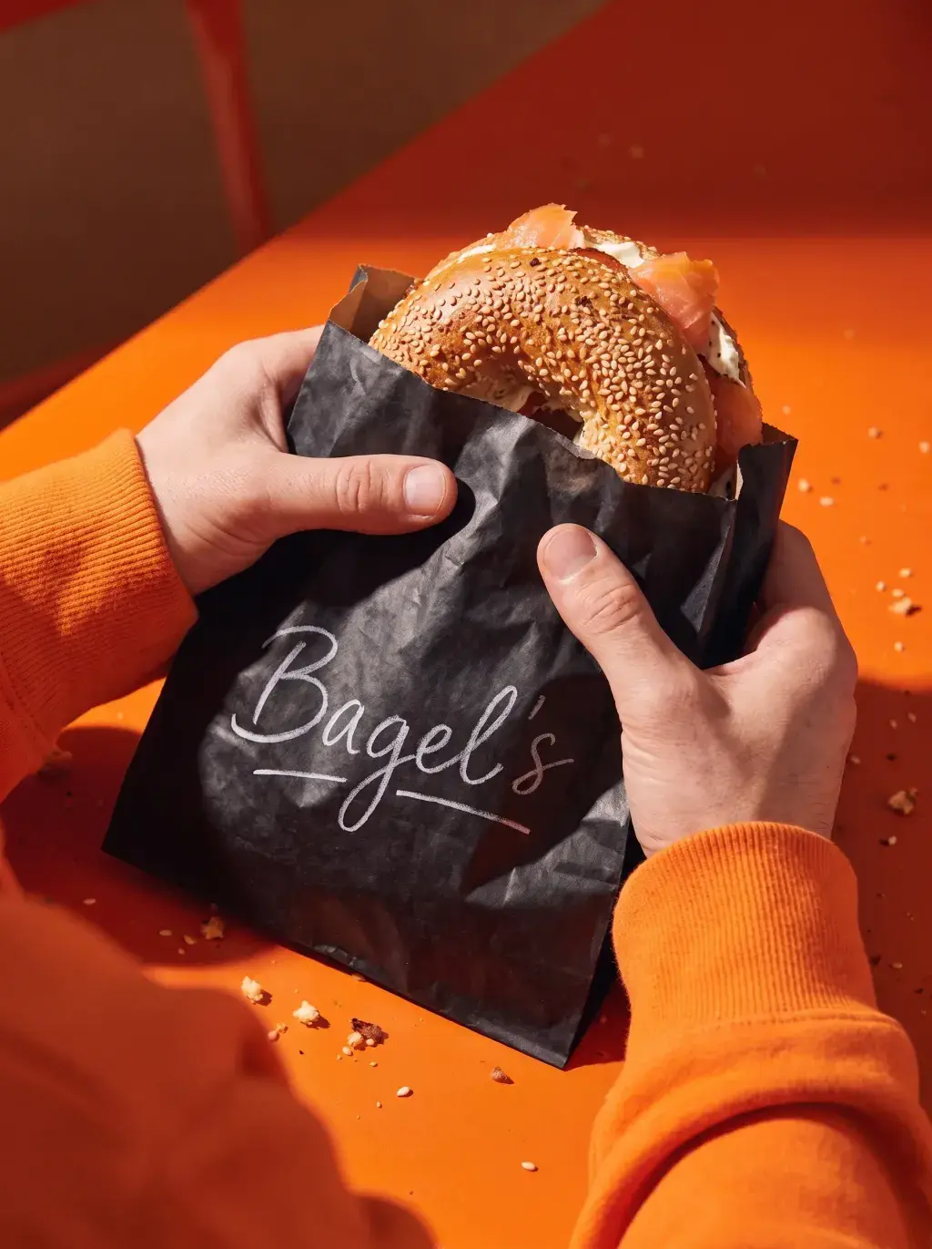 Mains en sweat orange tenant un sac Bagel's noir d'où dépasse un bagel saumon sur fond orange lumineux