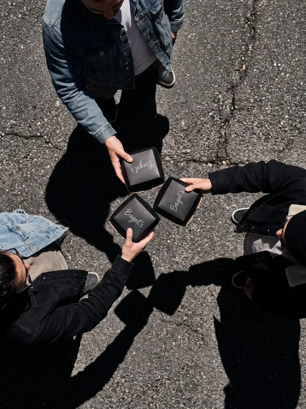 Vue aérienne de quatre mains tendant des boîtes Bagel's noires en cercle sur un sol en bitume ensoleillé