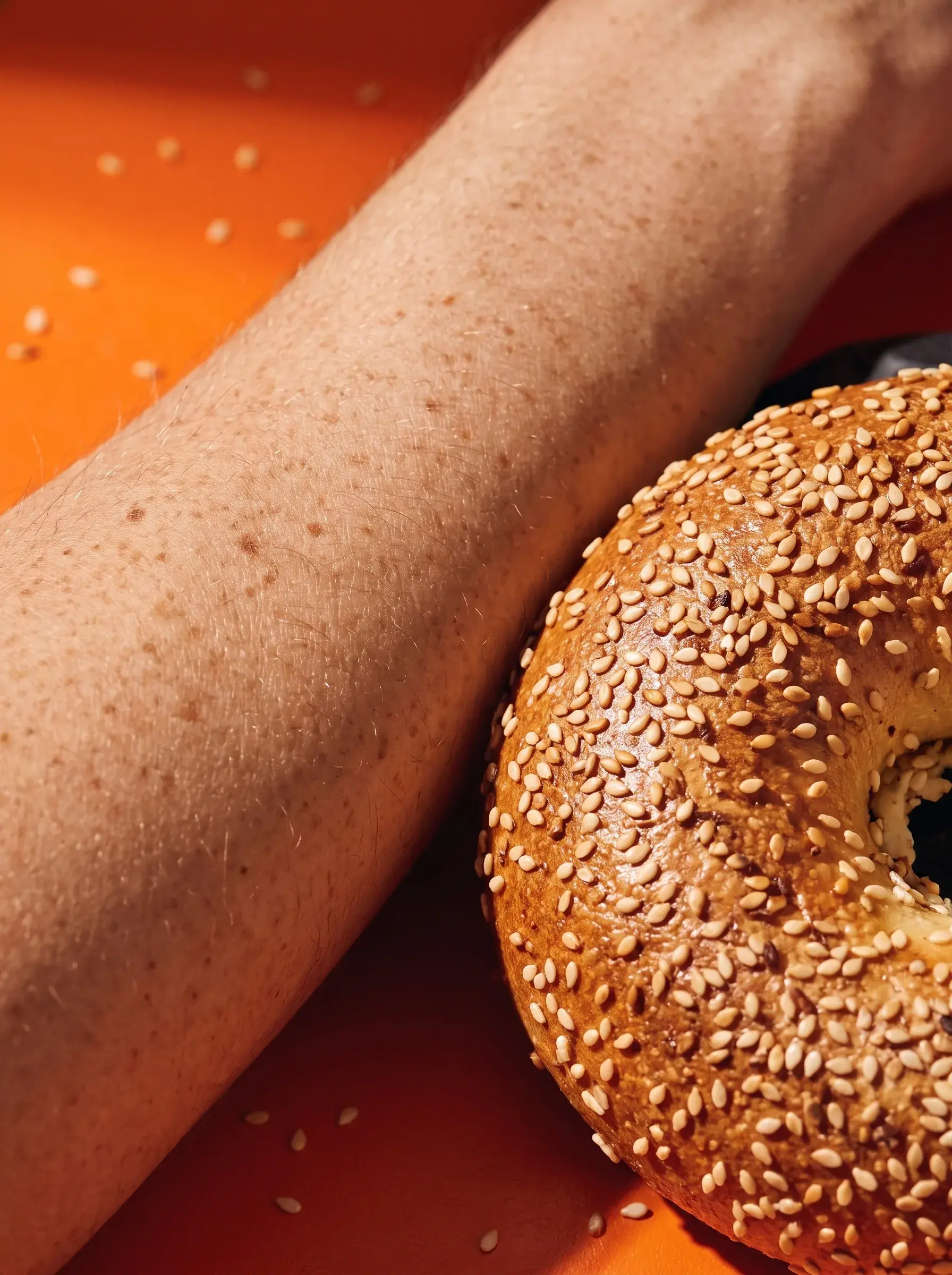 Gros plan d'un avant-bras parsemé de taches de rousseur posé contre un demi-bagel au sésame sur fond orange