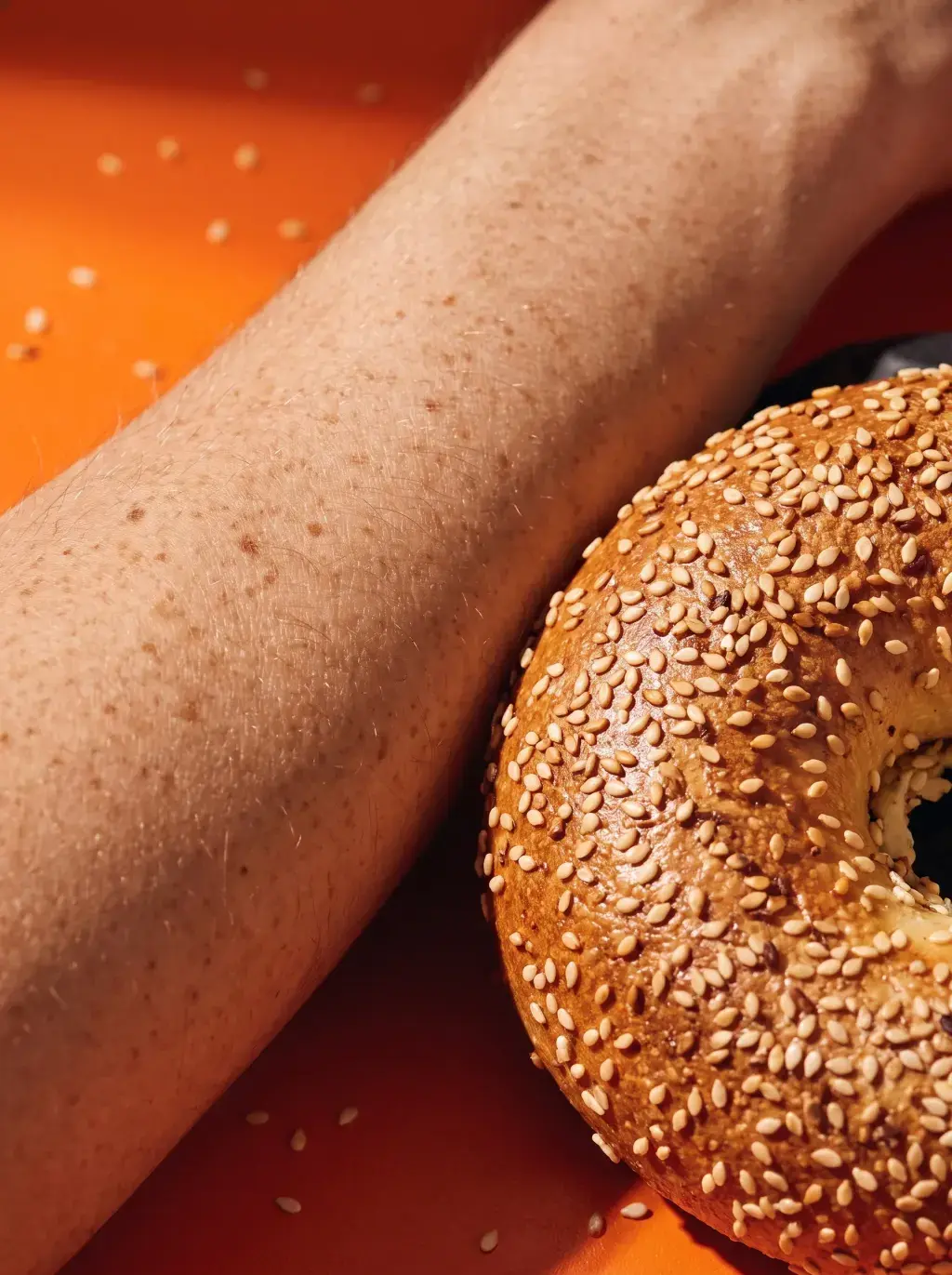 Gros plan d'un avant-bras parsemé de taches de rousseur posé contre un demi-bagel au sésame sur fond orange