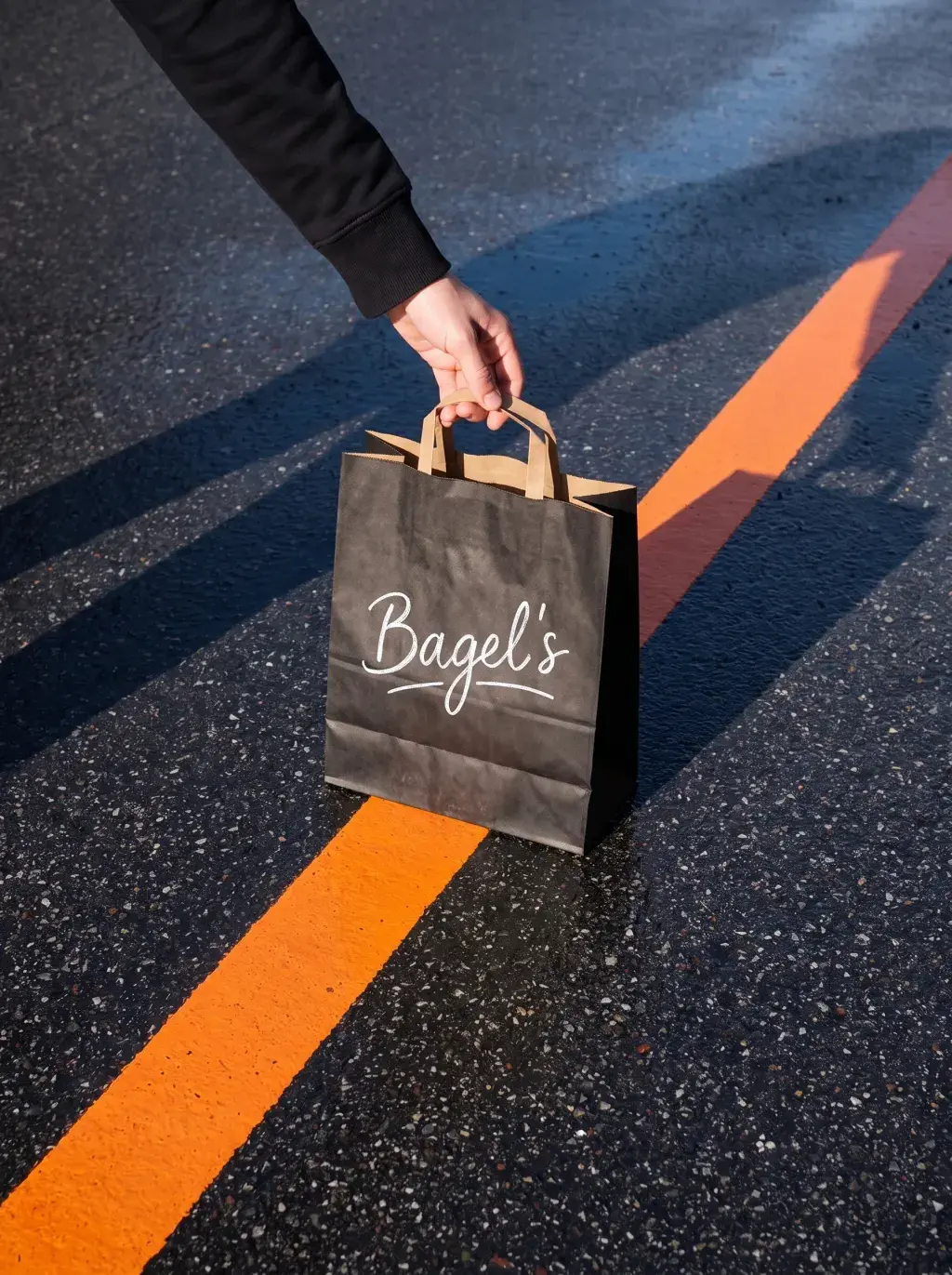 Main en sweat noir soulevant un sac Bagel's noir sur un sol en bitume avec une ligne orange peinte