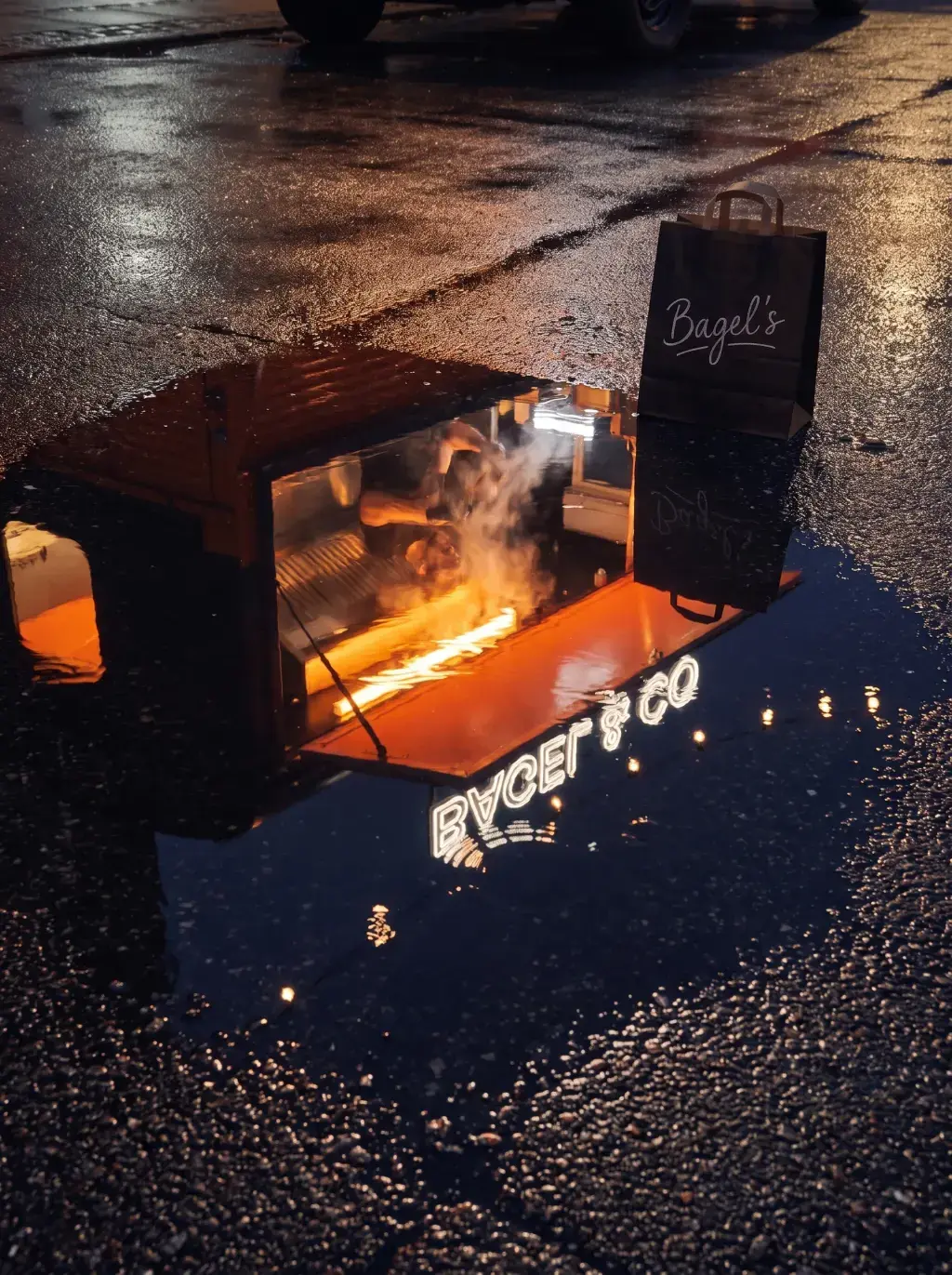 Reflet du food truck Bagel's orange et de l'enseigne BAGEL et CO dans une flaque de pluie nocturne avec sac et ardoise