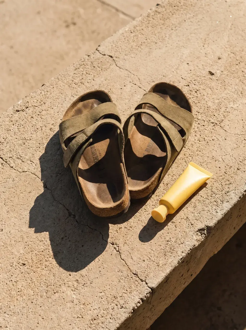 Sandales noires et tube cosmétique jaune posés sur une dalle de béton au soleil
