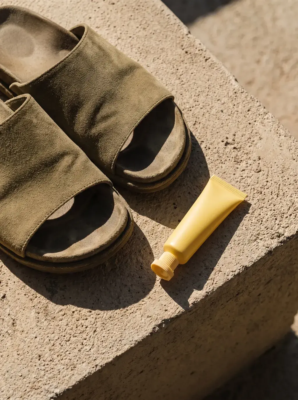 Sandales noires et tube jaune couchés sur une dalle minérale au soleil